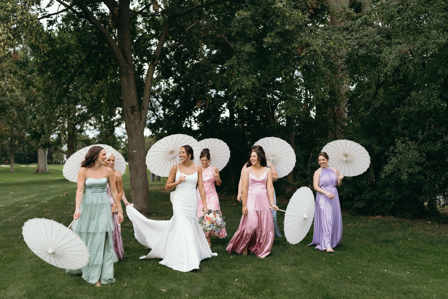 Bride and bridesmaids in colorful dresses holding white parasols on a green lawn with trees.