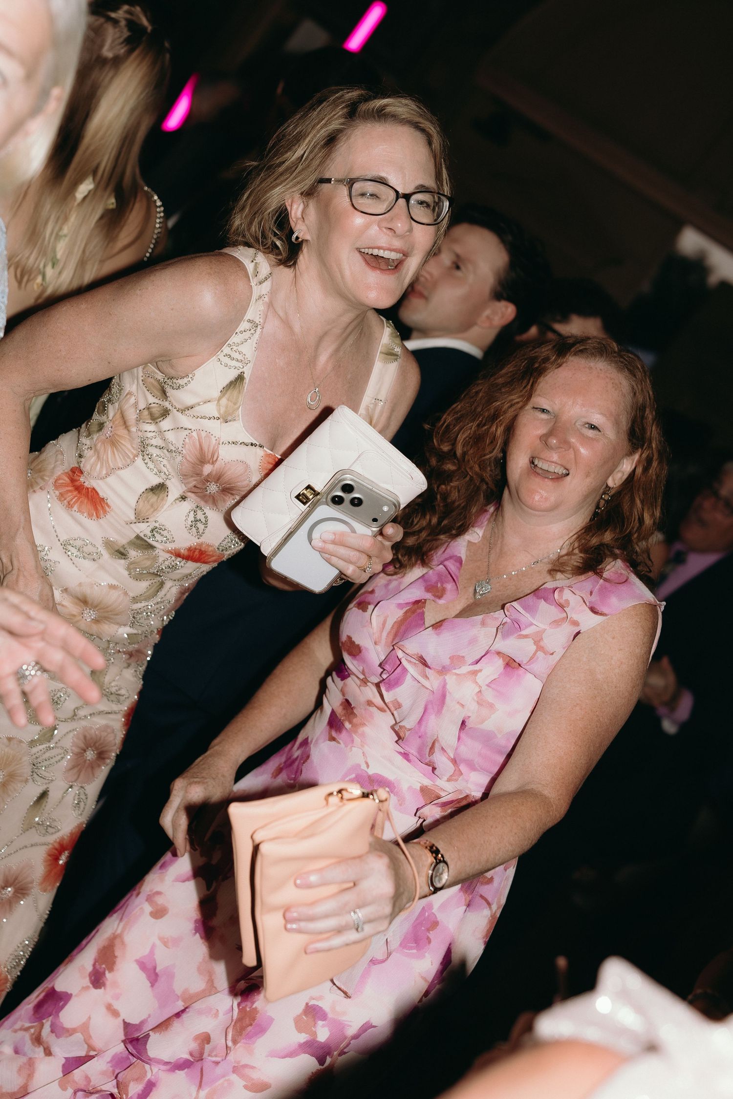 Two smiling women in floral dresses enjoying a fun moment together at a wedding reception.