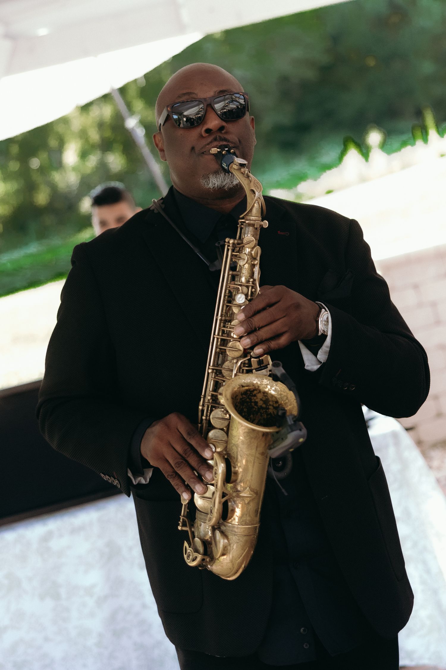 Musician in black suit and sunglasses playing a golden saxophone at an outdoor wedding reception.