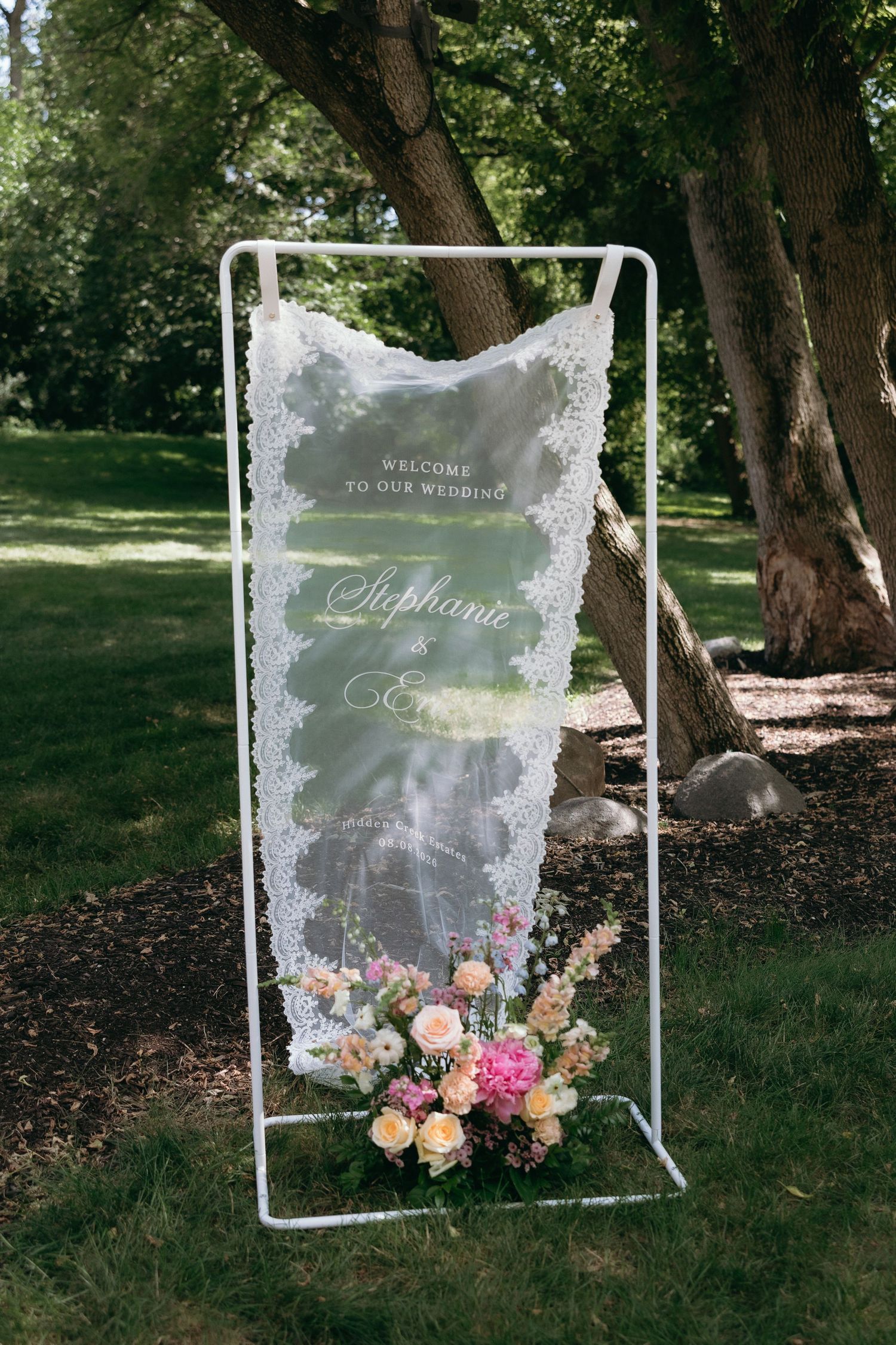 Elegant outdoor wedding welcome sign on white frame with lace fabric, Script names, and pink floral arrangement on green lawn.
