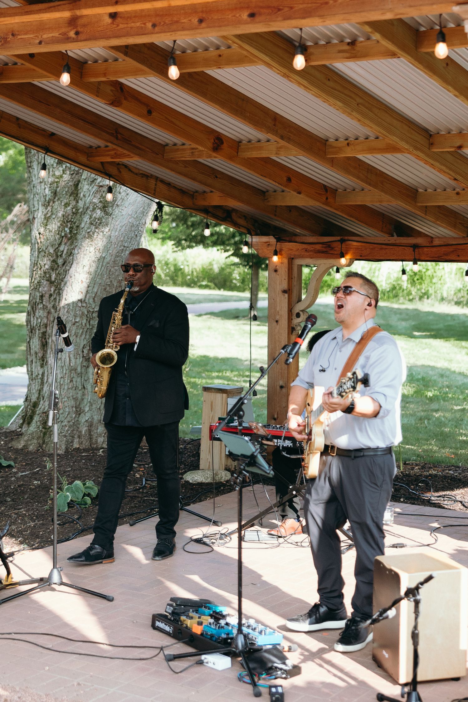 Live band with saxophonist and guitarist performing under wooden pavilion with string lights outdoors.