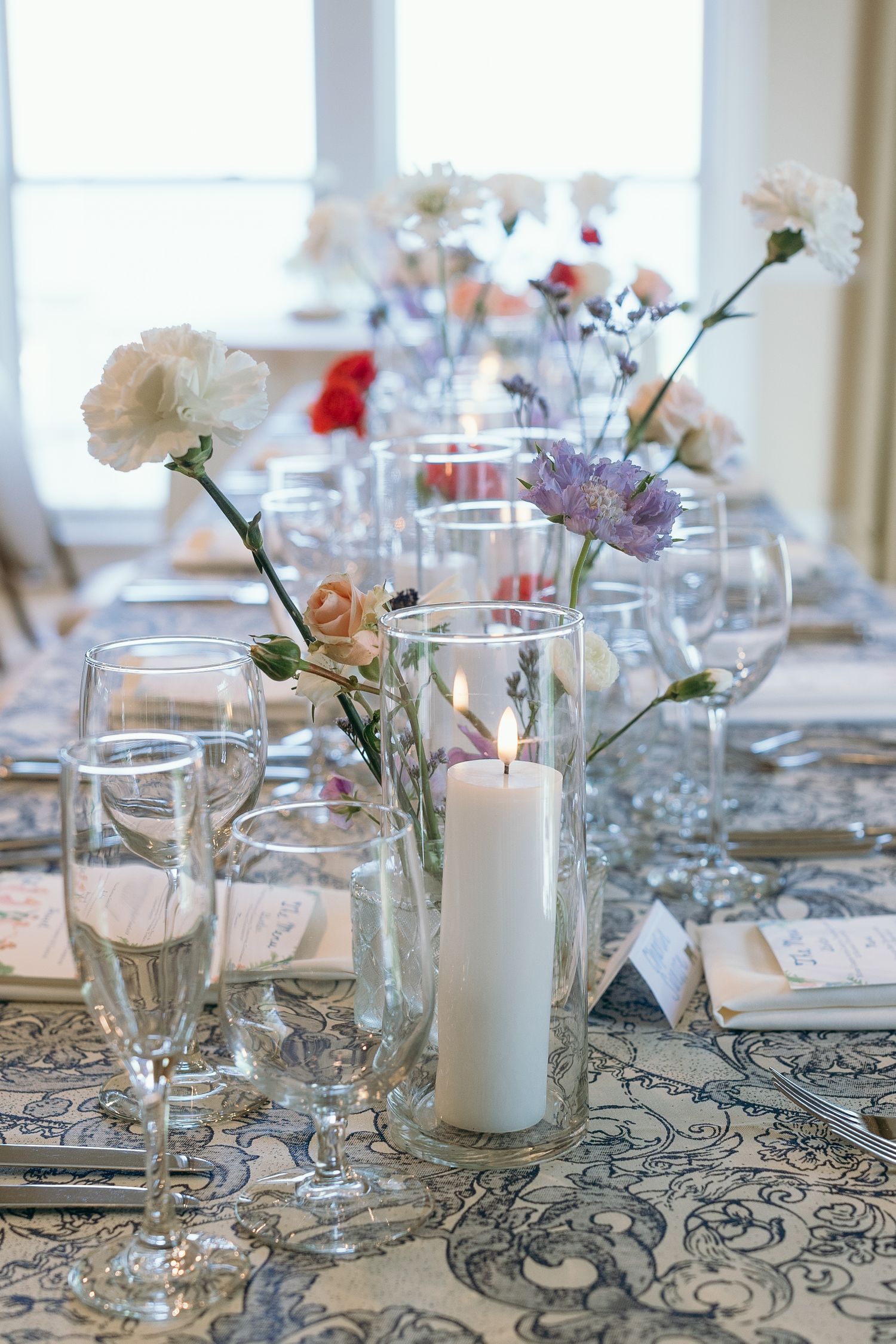 Elegant table setting with white candles, delicate flowers in glass vases, and patterned gray tablecloth for a wedding reception.