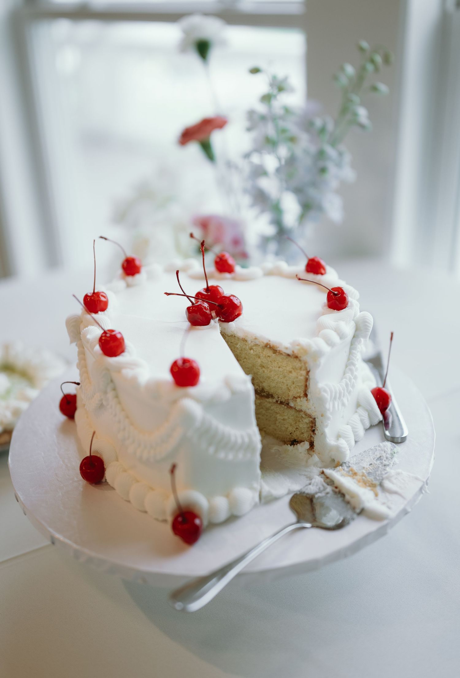 A white frosted layer cake topped with maraschino cherries sits on a cake stand with flowers in the background.