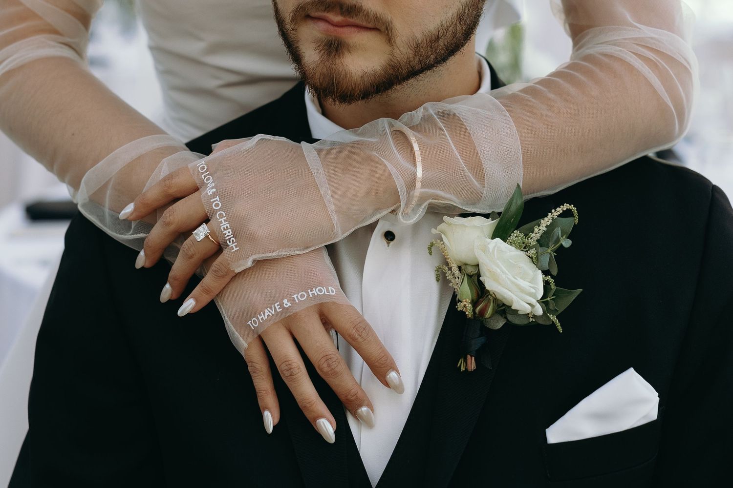 Close-up detail of hands with sheer sleeves and wedding rings against a black tuxedo.