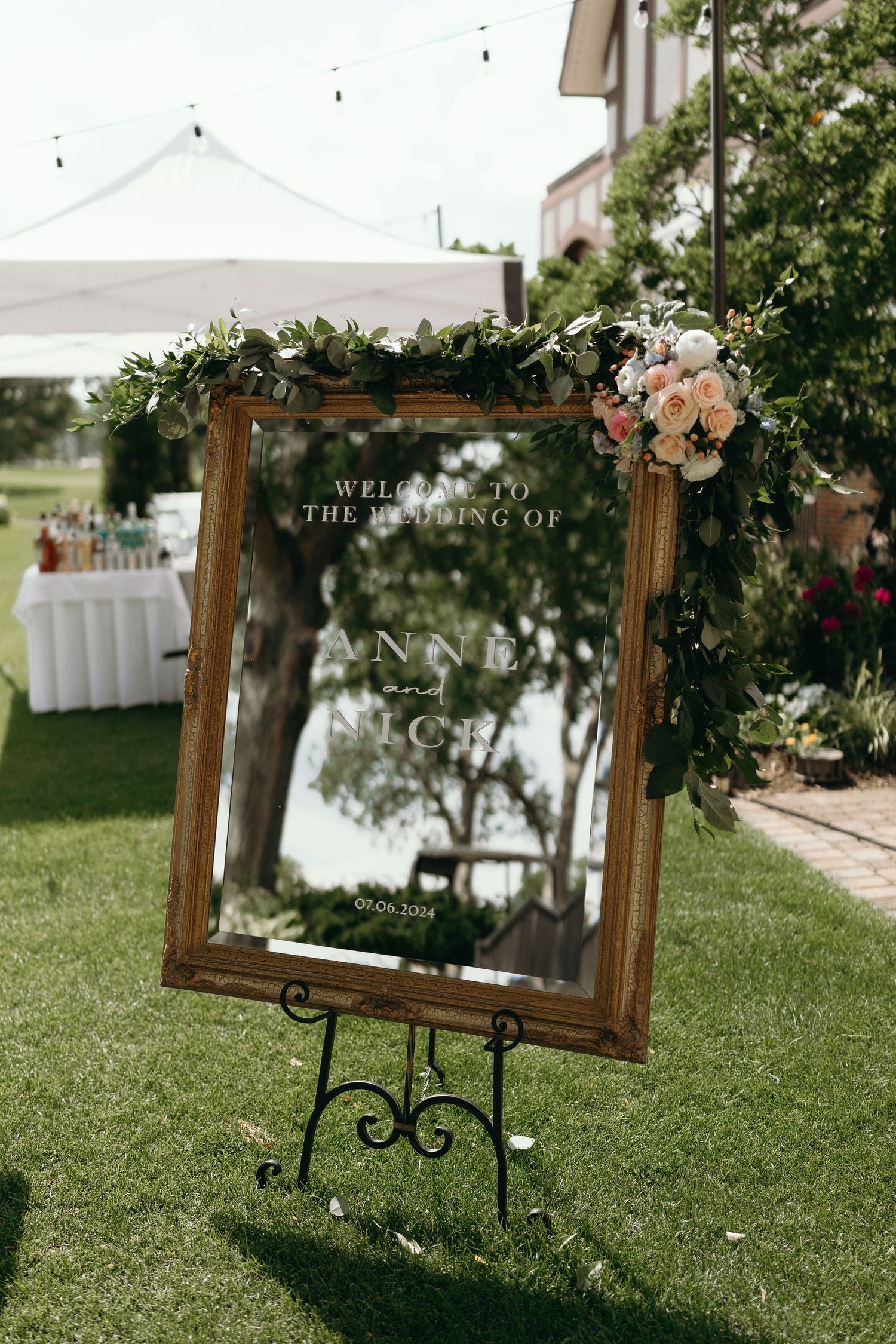 Ornate wooden frame decorated with flowers sits on grass at an outdoor wedding venue.
