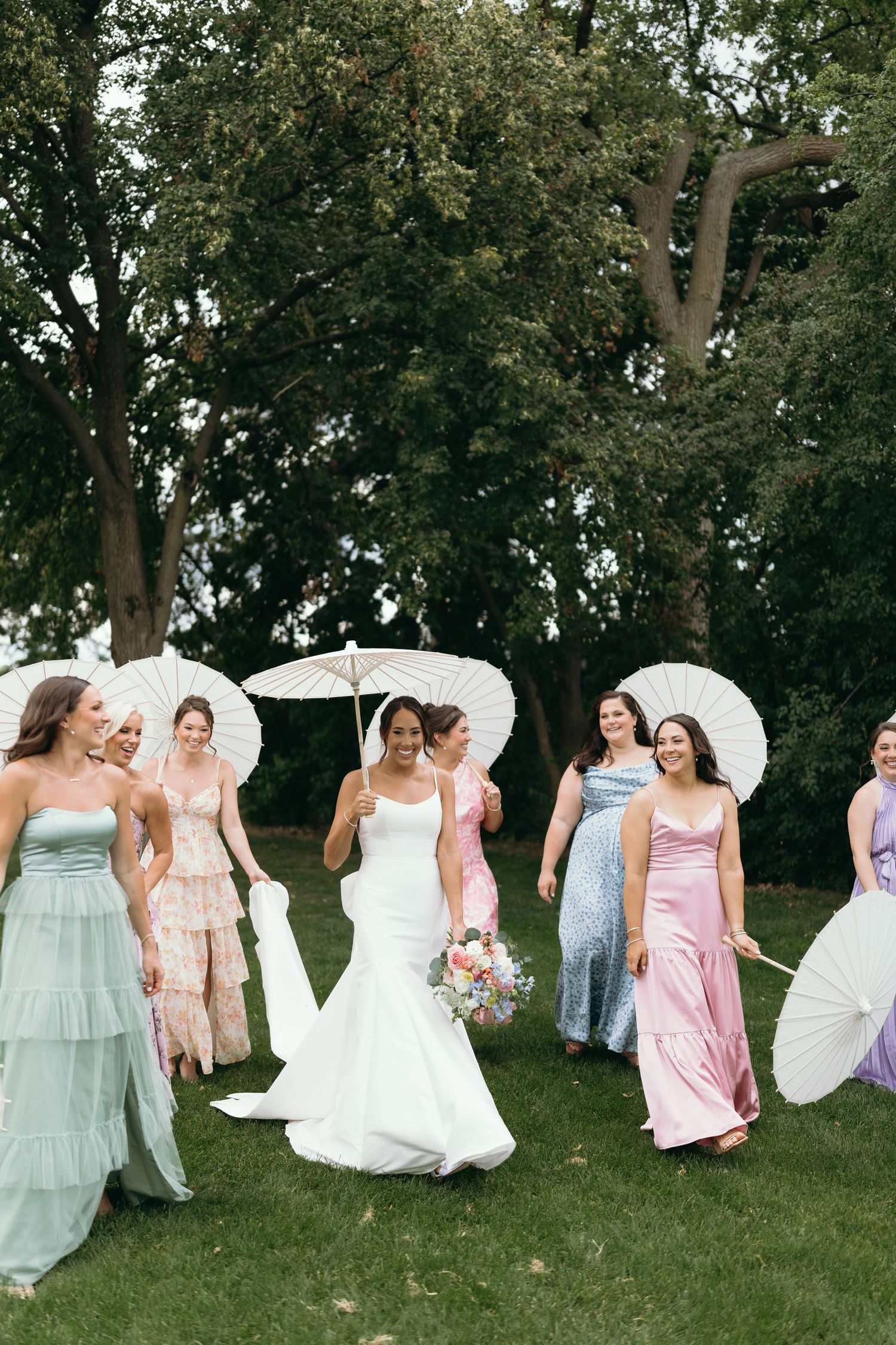 Wedding party posing with parasols under large trees in an outdoor garden setting.