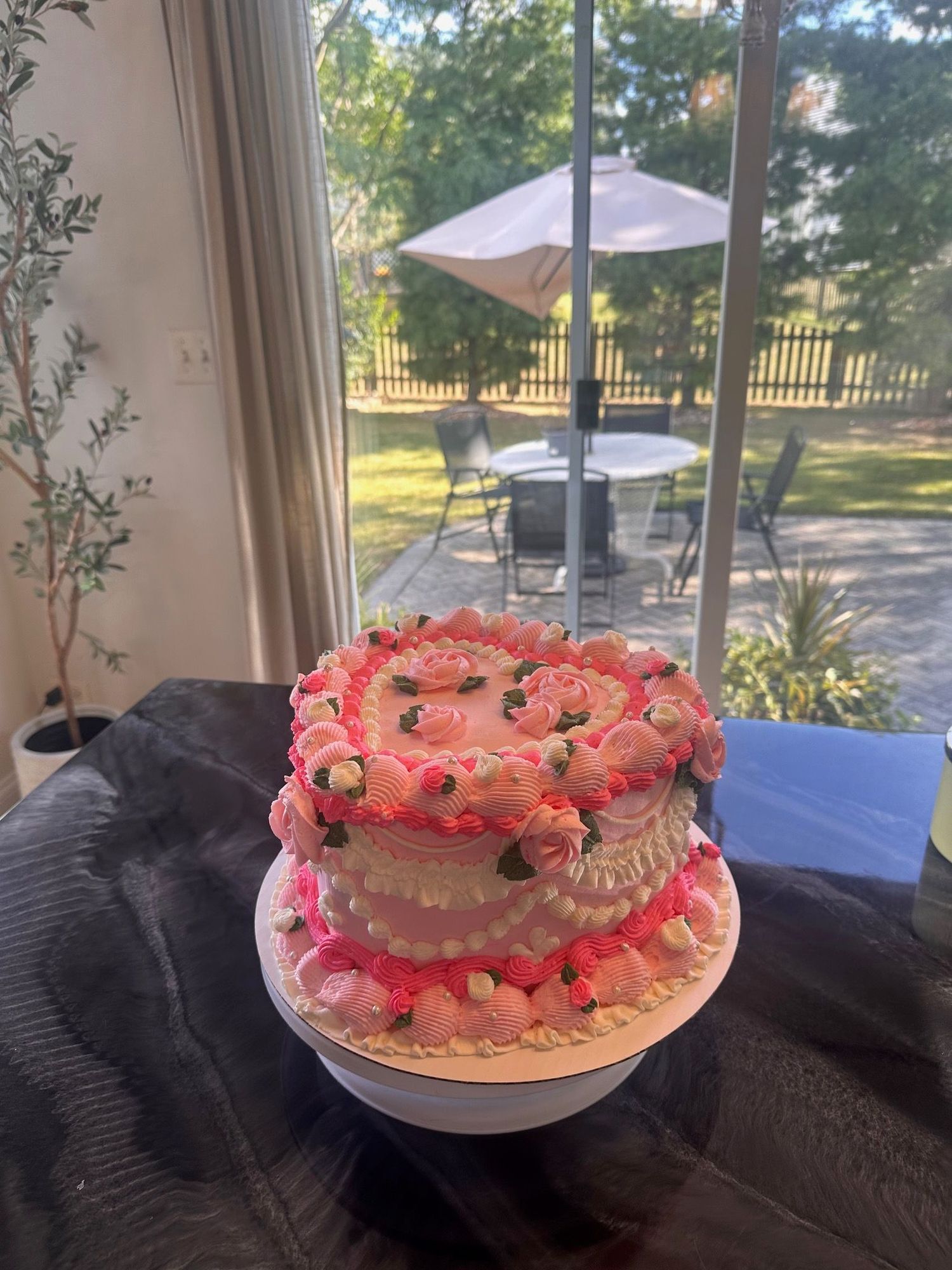 A pink frosted cake with decorative strawberries and whipped cream sits on a white plate near a window with outdoor view.