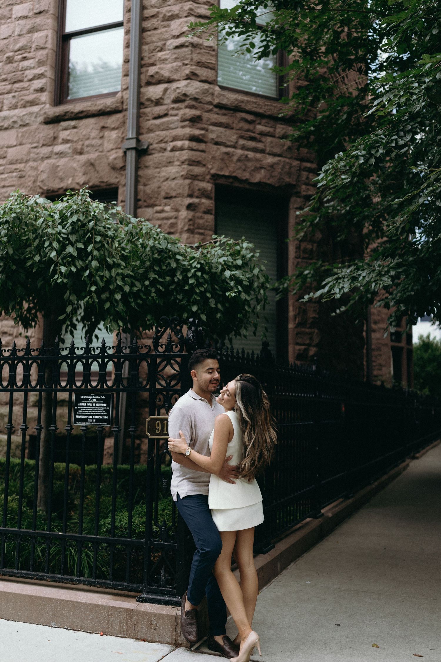 A couple embraces on a sidewalk outside a brownstone building with wrought iron fencing in an urban setting.