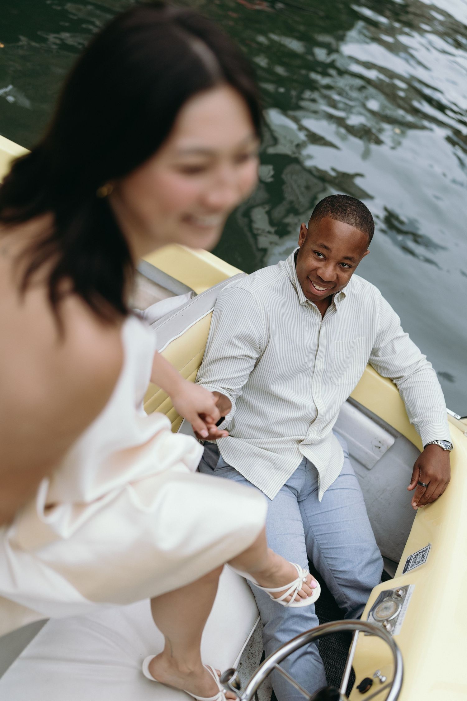 A smiling couple enjoys a romantic boat ride on calm water, with a woman in white and a man in a light shirt.