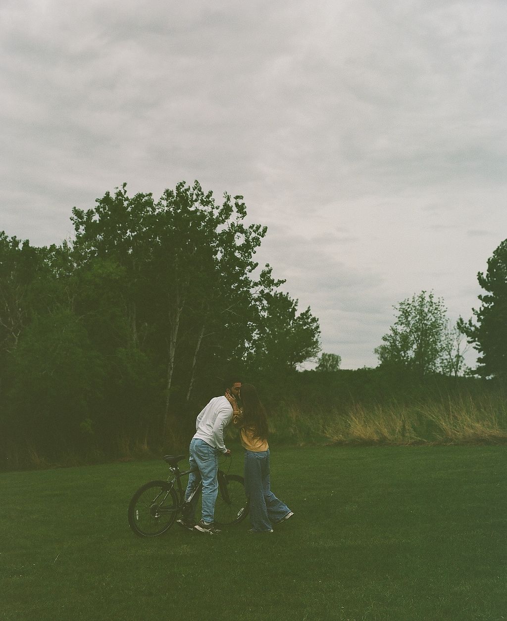 Couple sharing a romantic moment beside a bicycle on a lush green lawn with trees in the background.