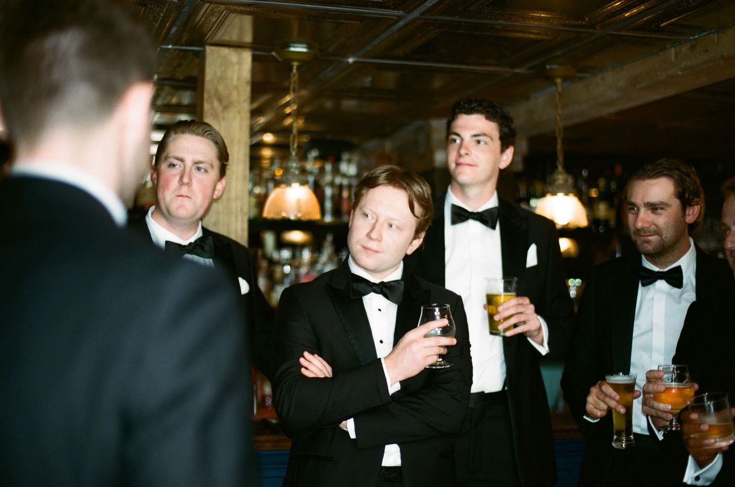 Men in black tuxedos and bow ties socializing at an elegant bar, holding cocktails at a formal evening event.
