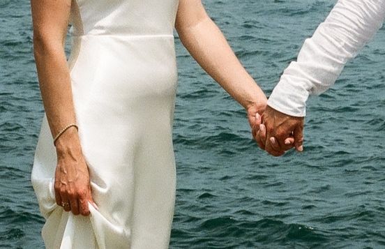 Interracial couple holding hands in wedding attire with ocean background, bride in white dress and groom in white suit.