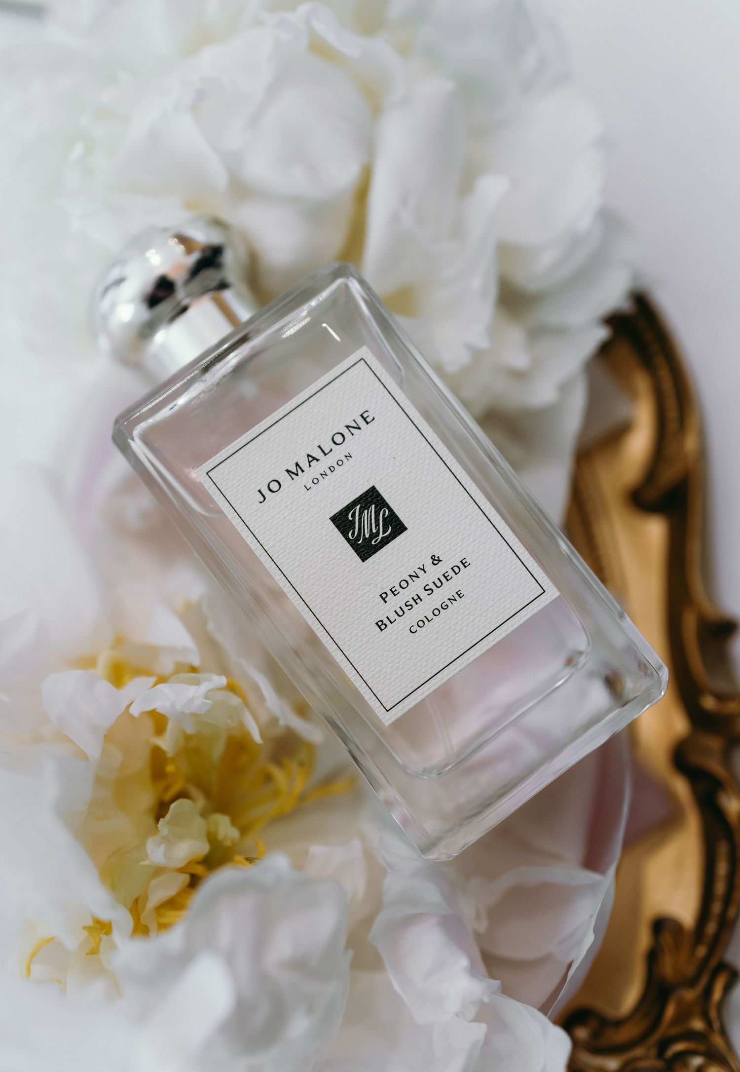 A luxurious Jo Malone perfume bottle with white peony flowers and ornate golden decor against a soft white background.