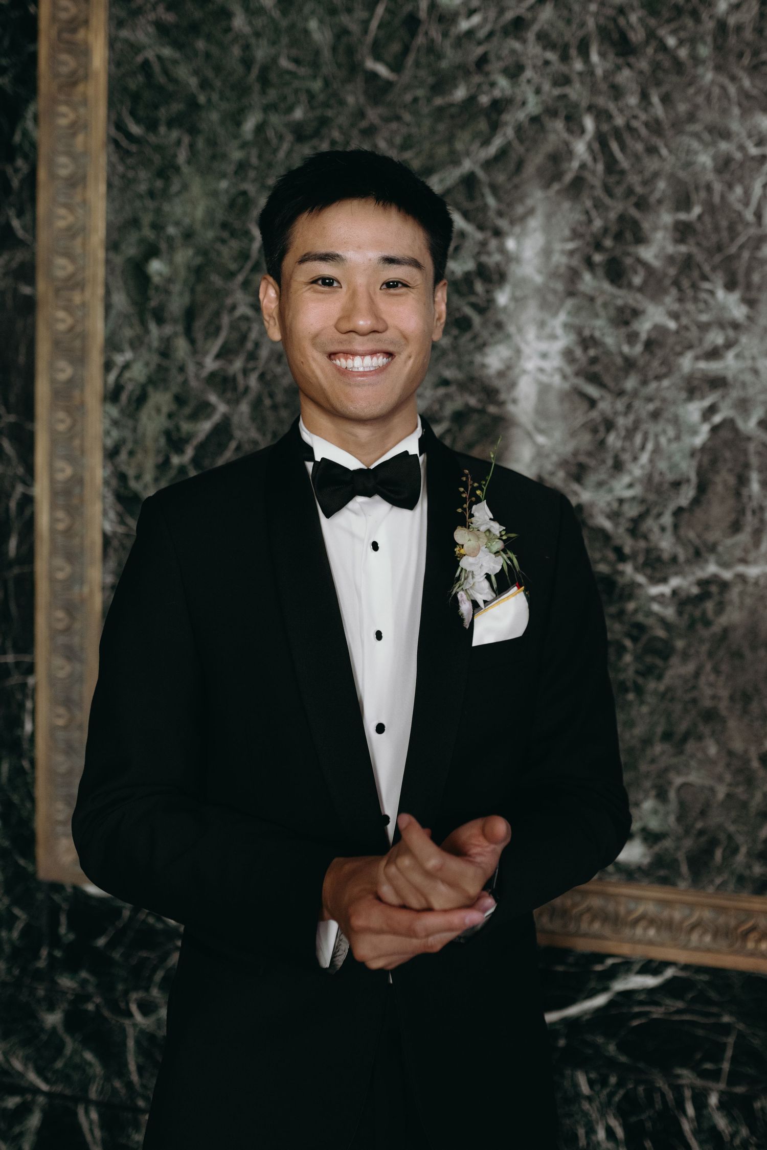 Formal portrait in a black tuxedo with white boutonniere against dark marble background.