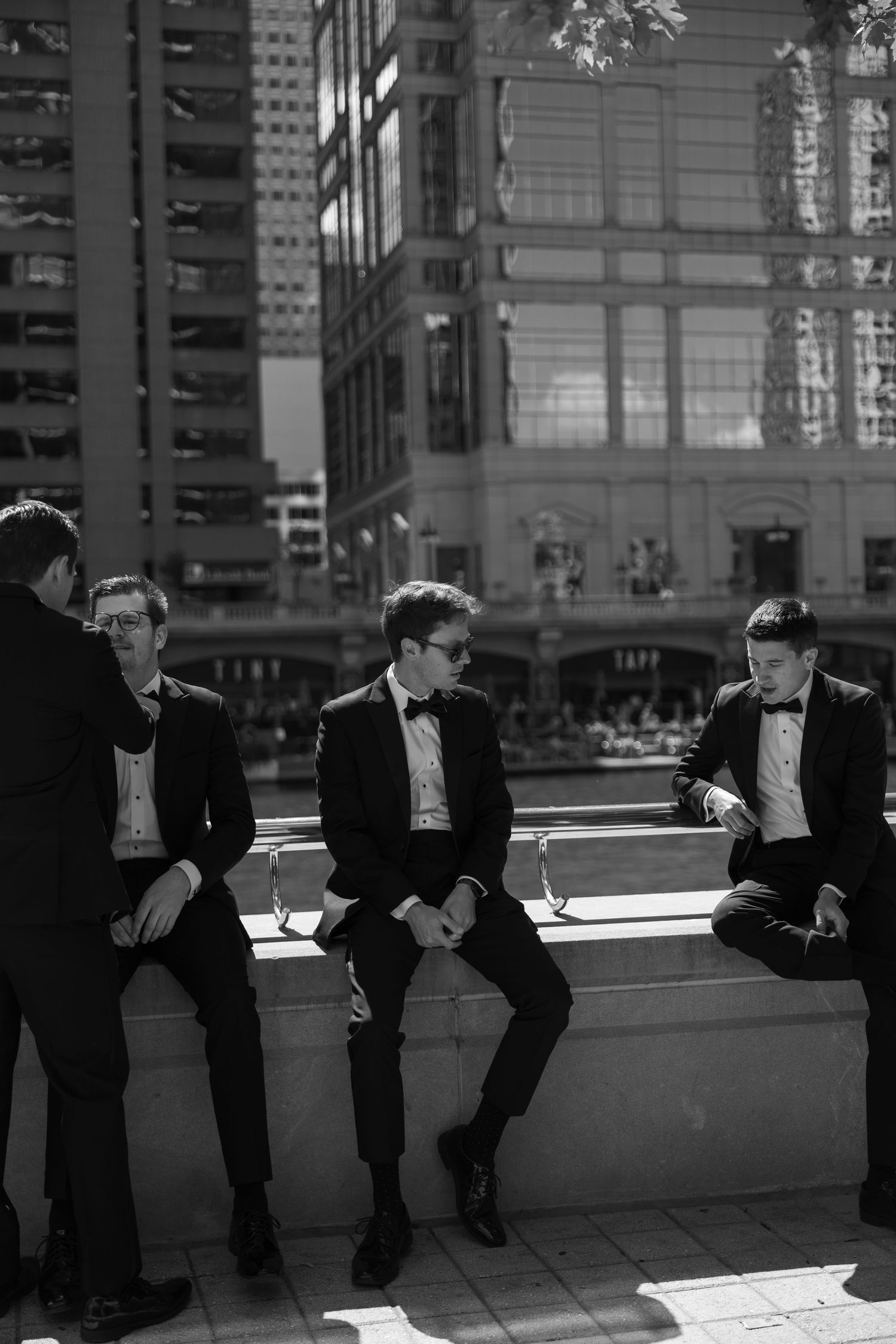 Groomsmen in black tuxedos sitting casually on urban ledge in black and white photography.