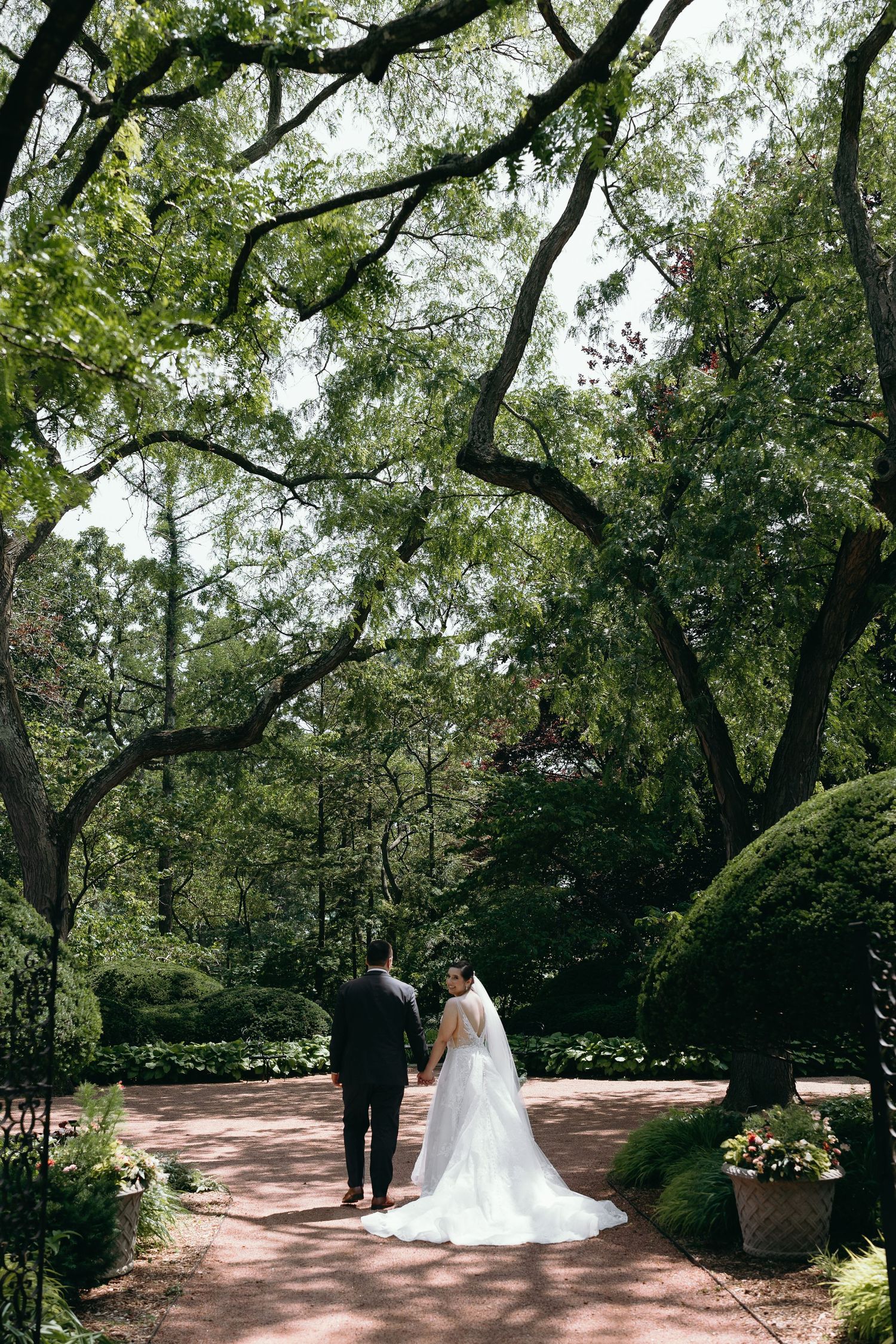 A couple walking down a tree-lined garden path in their wedding attire.