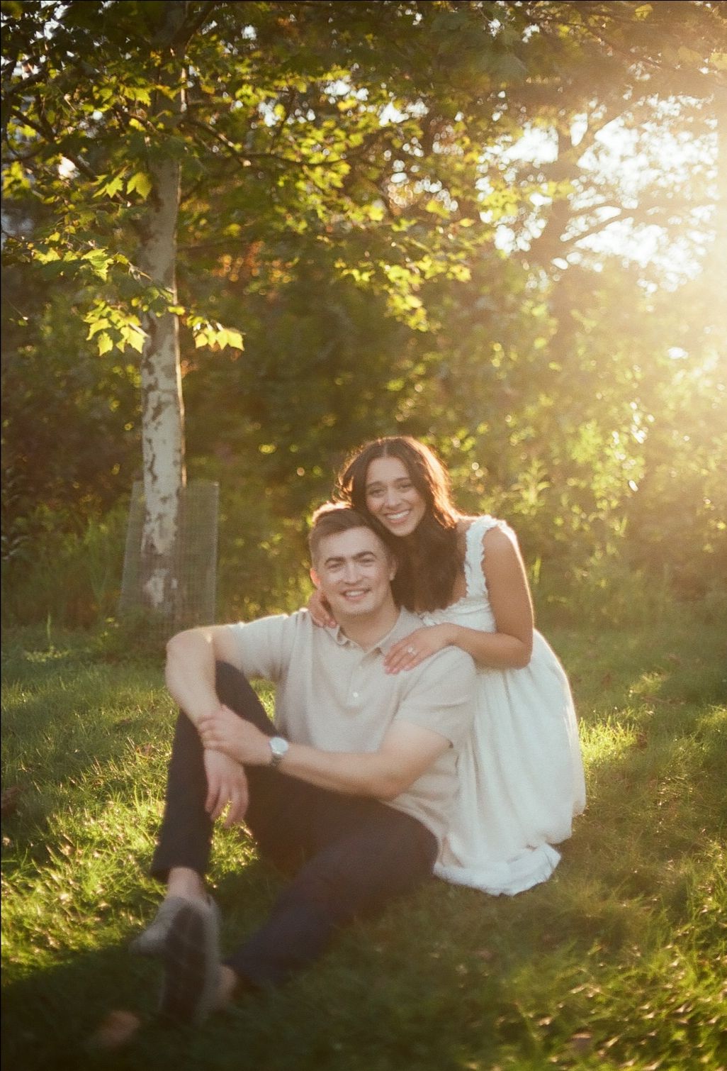 A couple sits together in a sunlit forest clearing during golden hour, her in a white dress and him in casual attire.