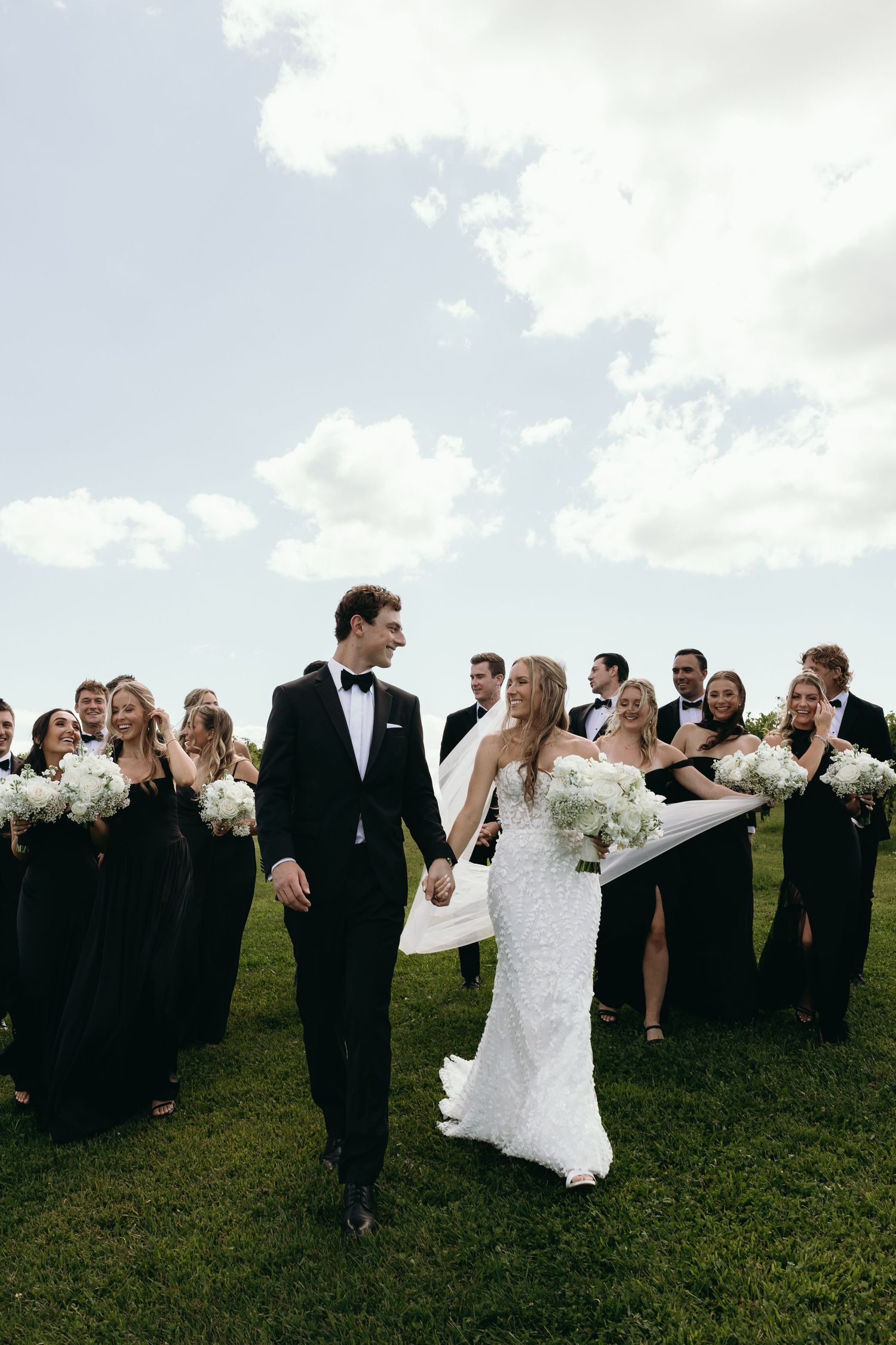 Wedding party in black and white attire walks across grassy hilltop under cloudy sky during outdoor celebration.