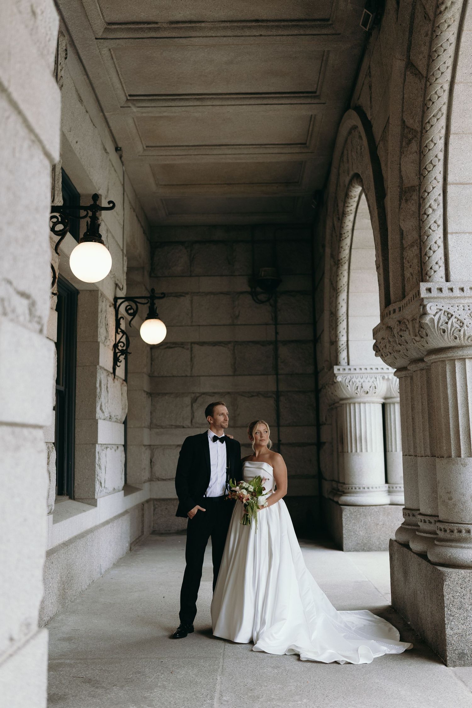 A couple poses in an elegant colonnade hallway with white stone arches and pillars for wedding photography.