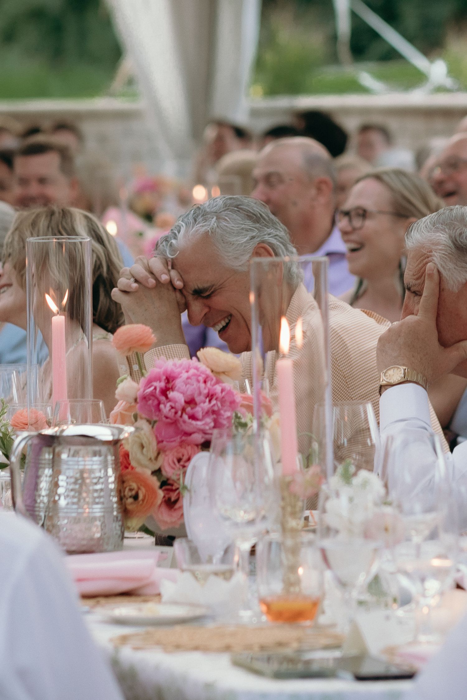 Wedding guests laughing joyfully at a beautifully decorated reception table with pink floral centerpieces.