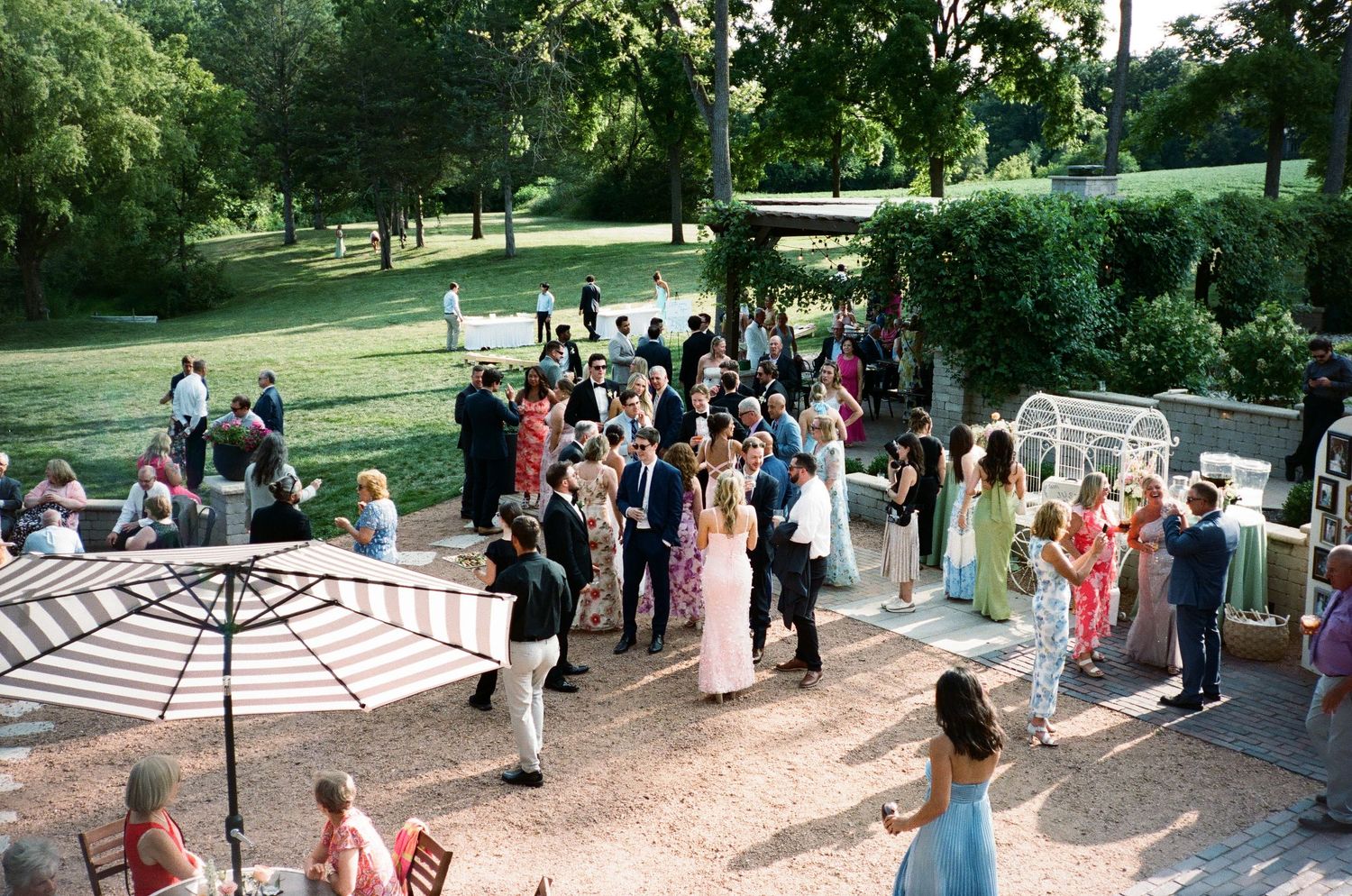 Elegant outdoor wedding reception on a patio with guests mingling, surrounded by lush green trees and manicured lawns.
