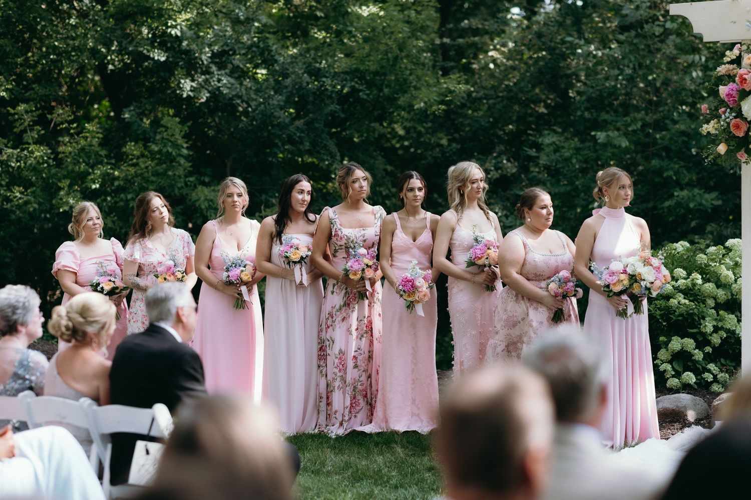 Nine bridesmaids in pink dresses holding floral bouquets stand together at an outdoor wedding ceremony.