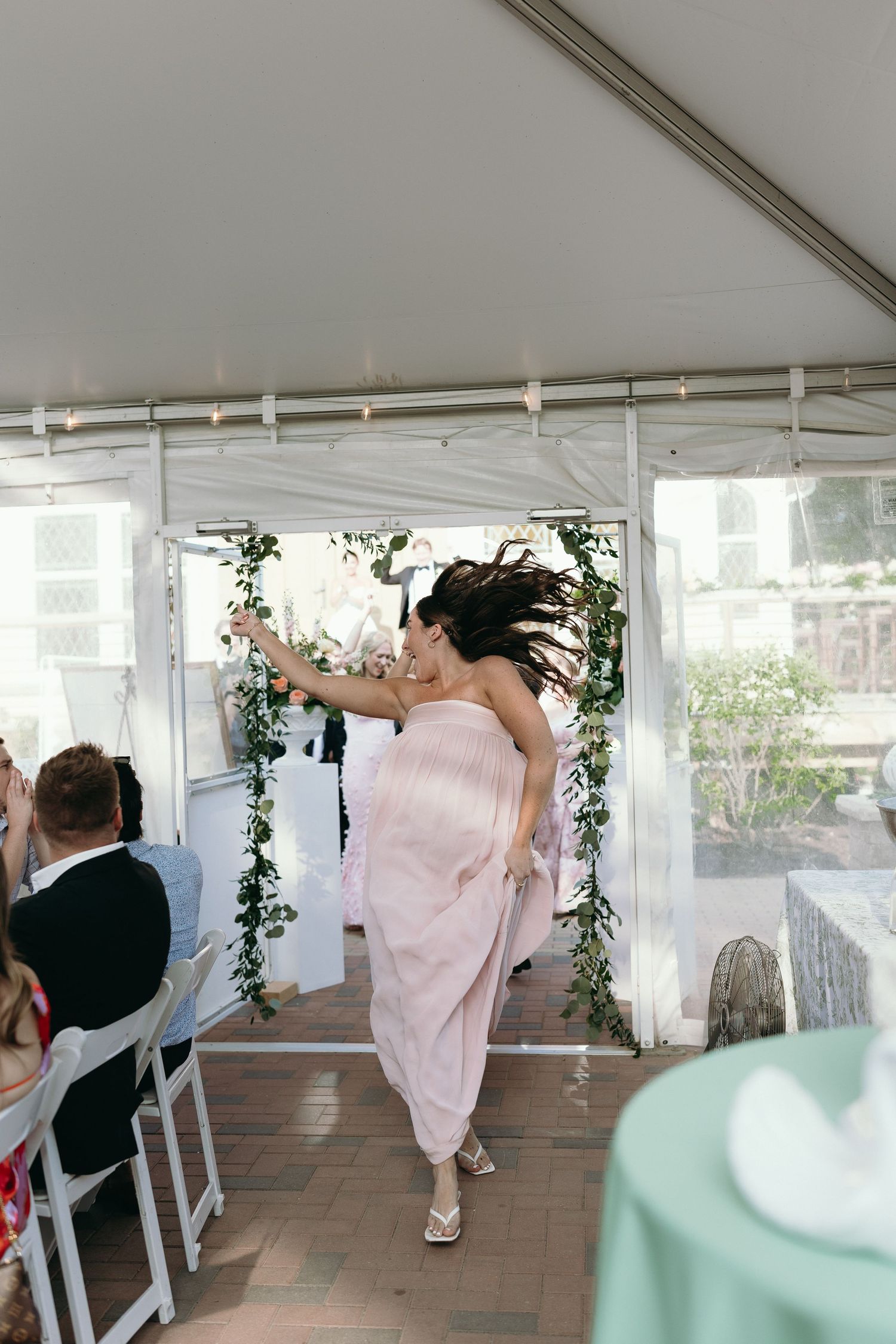 Bridesmaid in pink dress making energetic entrance at outdoor wedding reception under white tent.