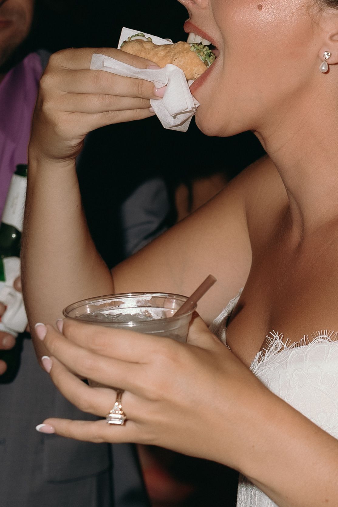 Bride in white dress eating burger and holding cocktail wearing diamond engagement ring.