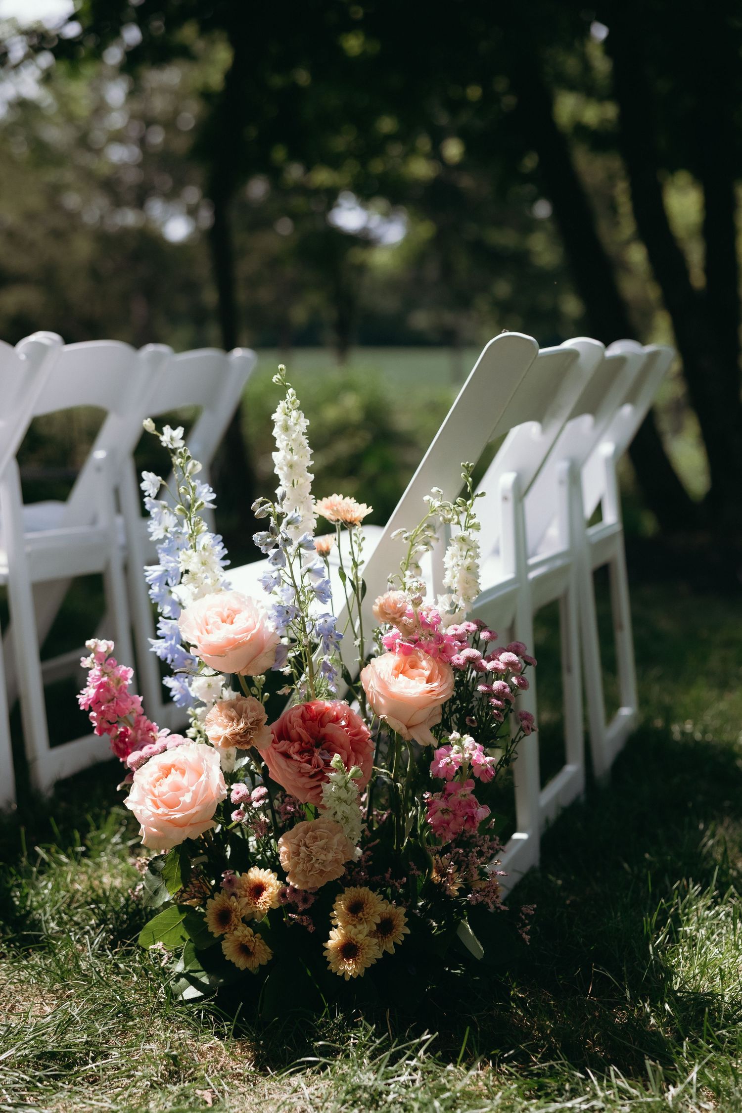 Colorful wedding aisle floral arrangement with pink, peach, and blue flowers beside white folding chairs on green grass.