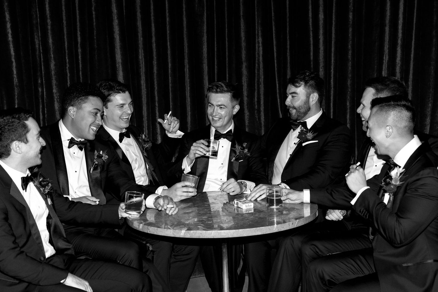 Seven groomsmen in tuxedos laugh together around a small round table in a black and white wedding photo.