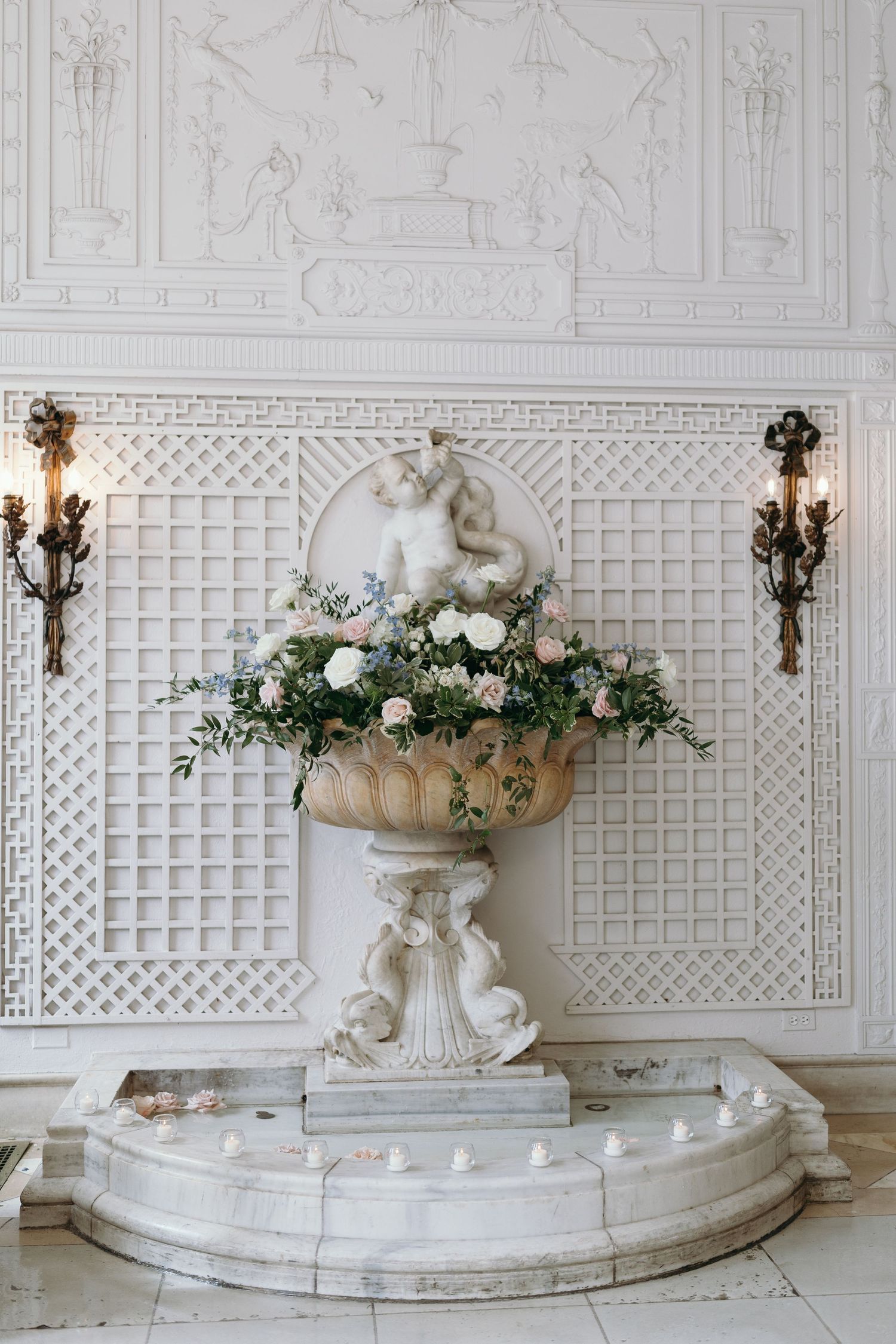 An ornate white stone planter filled with white roses against a latticed wall with sconces.