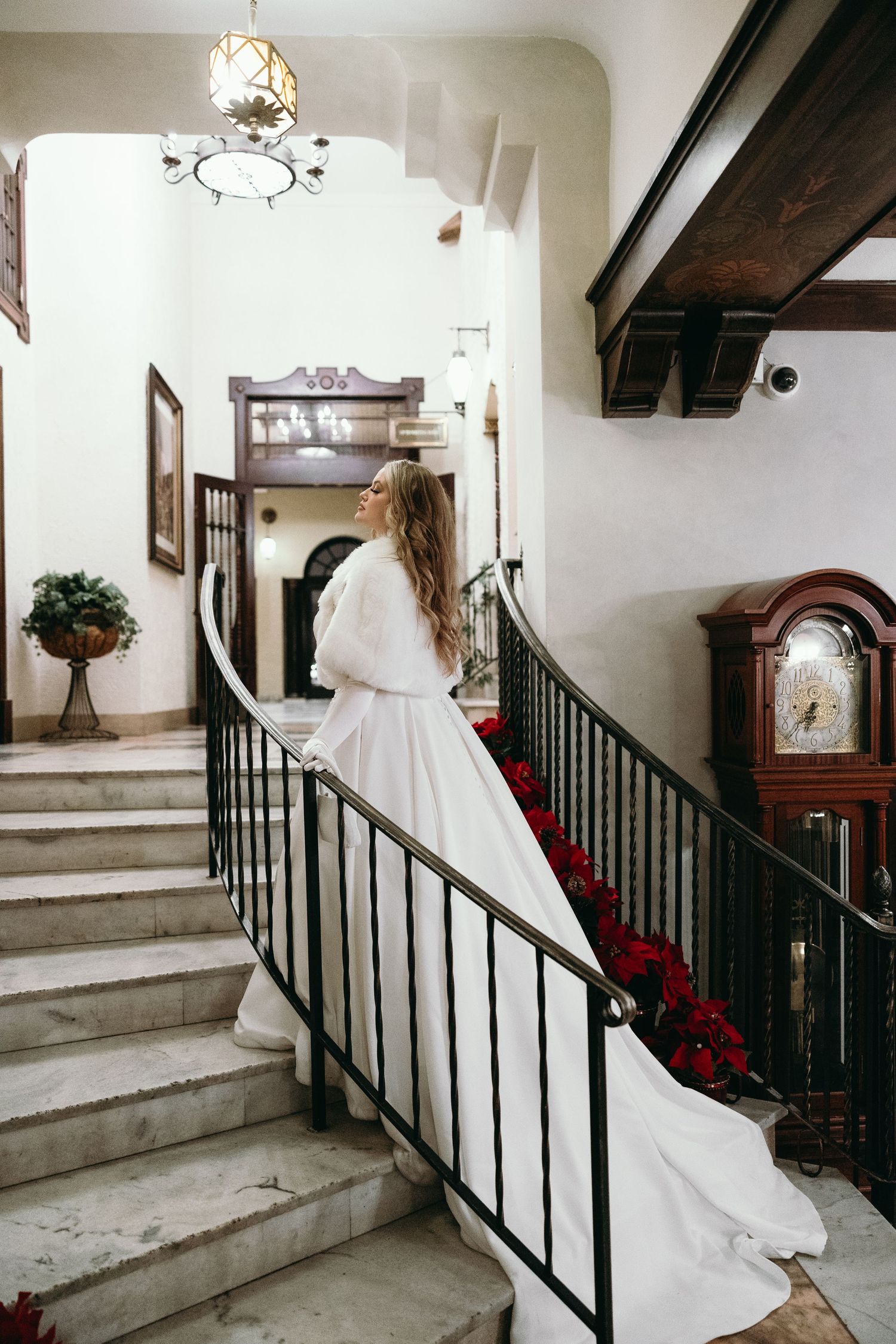 White wedding dress flowing down elegant staircase with red flowers and dark railings.