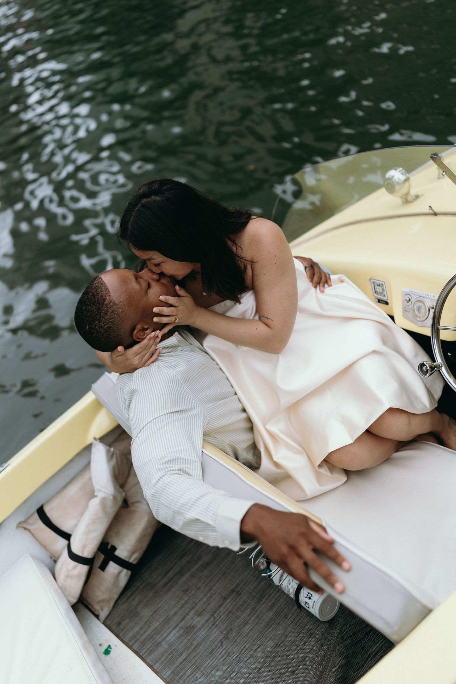 Romantic couple sharing a kiss on a vintage boat on the water, dressed in elegant white wedding attire.