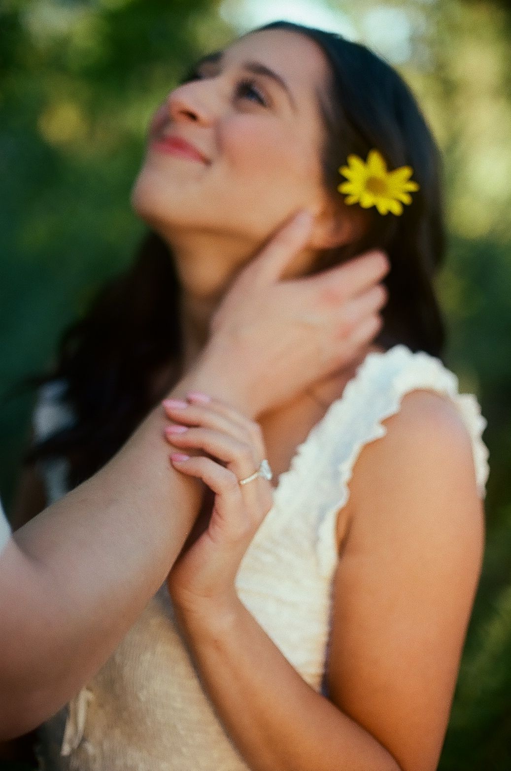 A figure in a white lace dress poses outdoors with a yellow sunflower against a soft bokeh background.