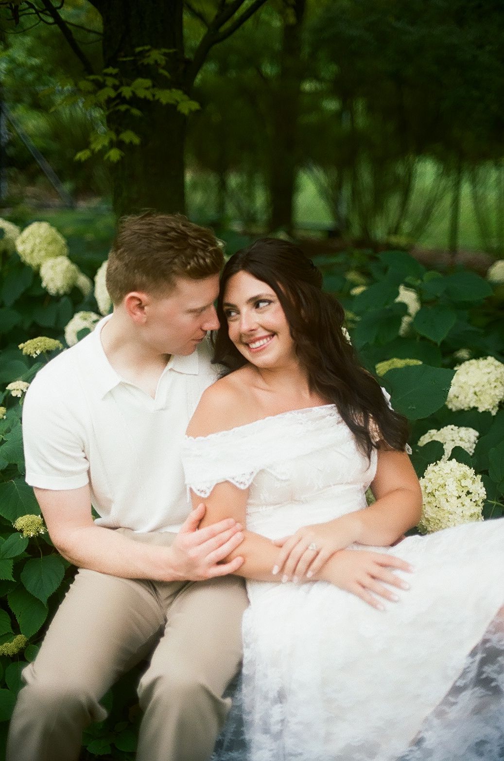 A romantic series of moments between a couple in white sitting together outdoors surrounded by green foliage and flowers.