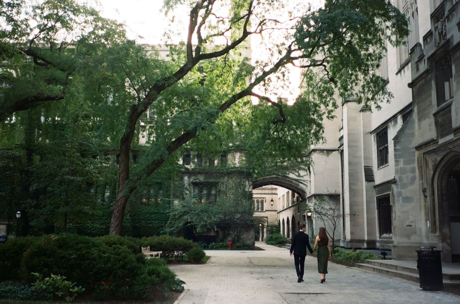 A couple walking along a tree-lined path near historic university buildings. Chicago Film Wedding Photographer