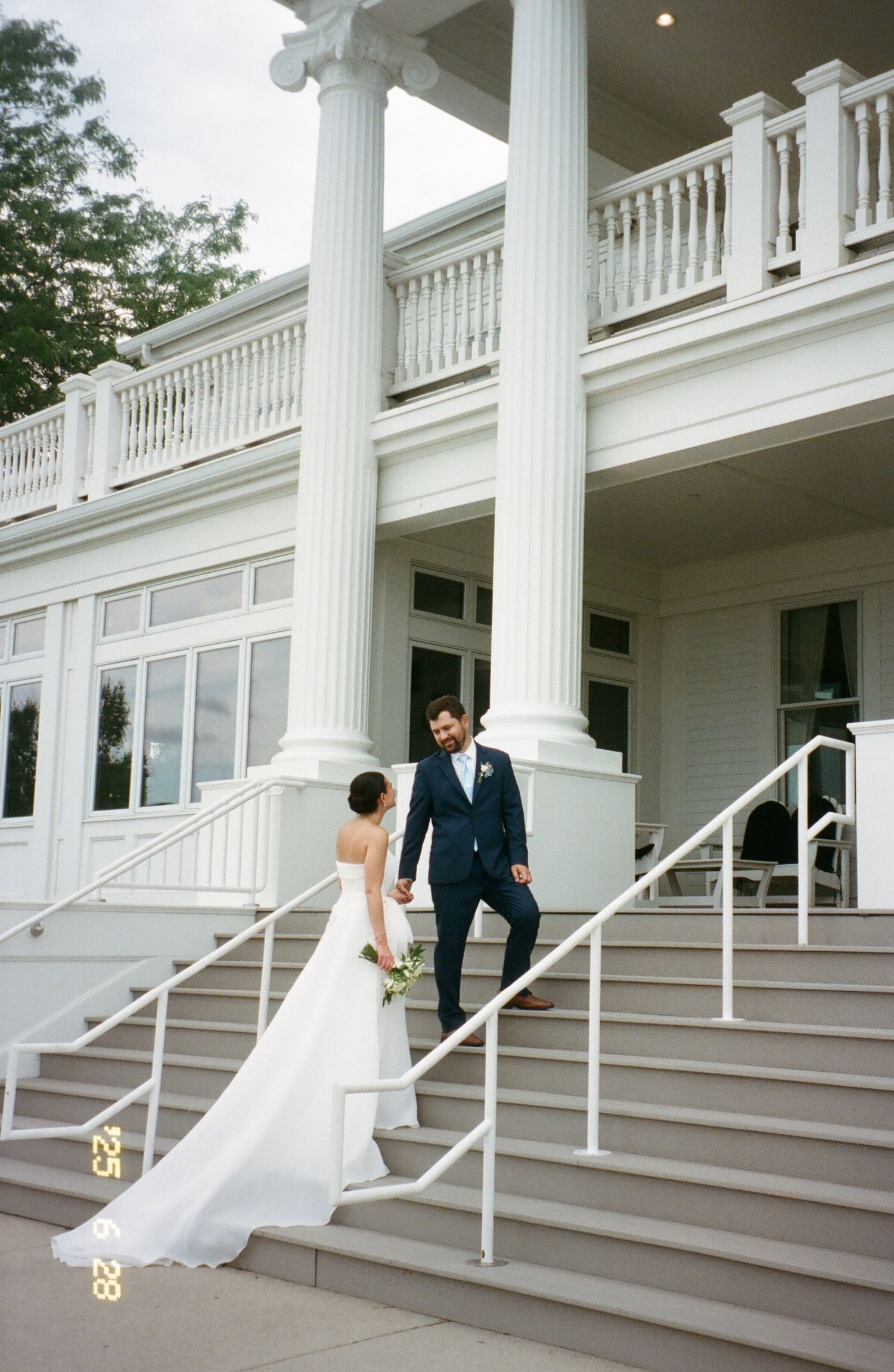 A couple descends white stairs at a classic colonial style venue with grand columns and railings on their wedding day.