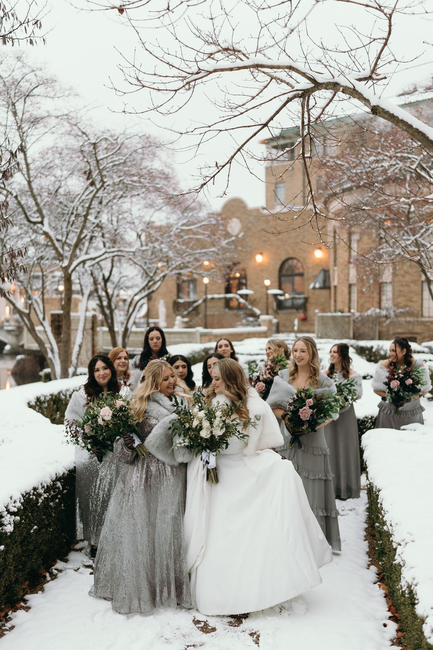 Bridal party with grey dresses and bouquets poses in snowy winter garden setting.