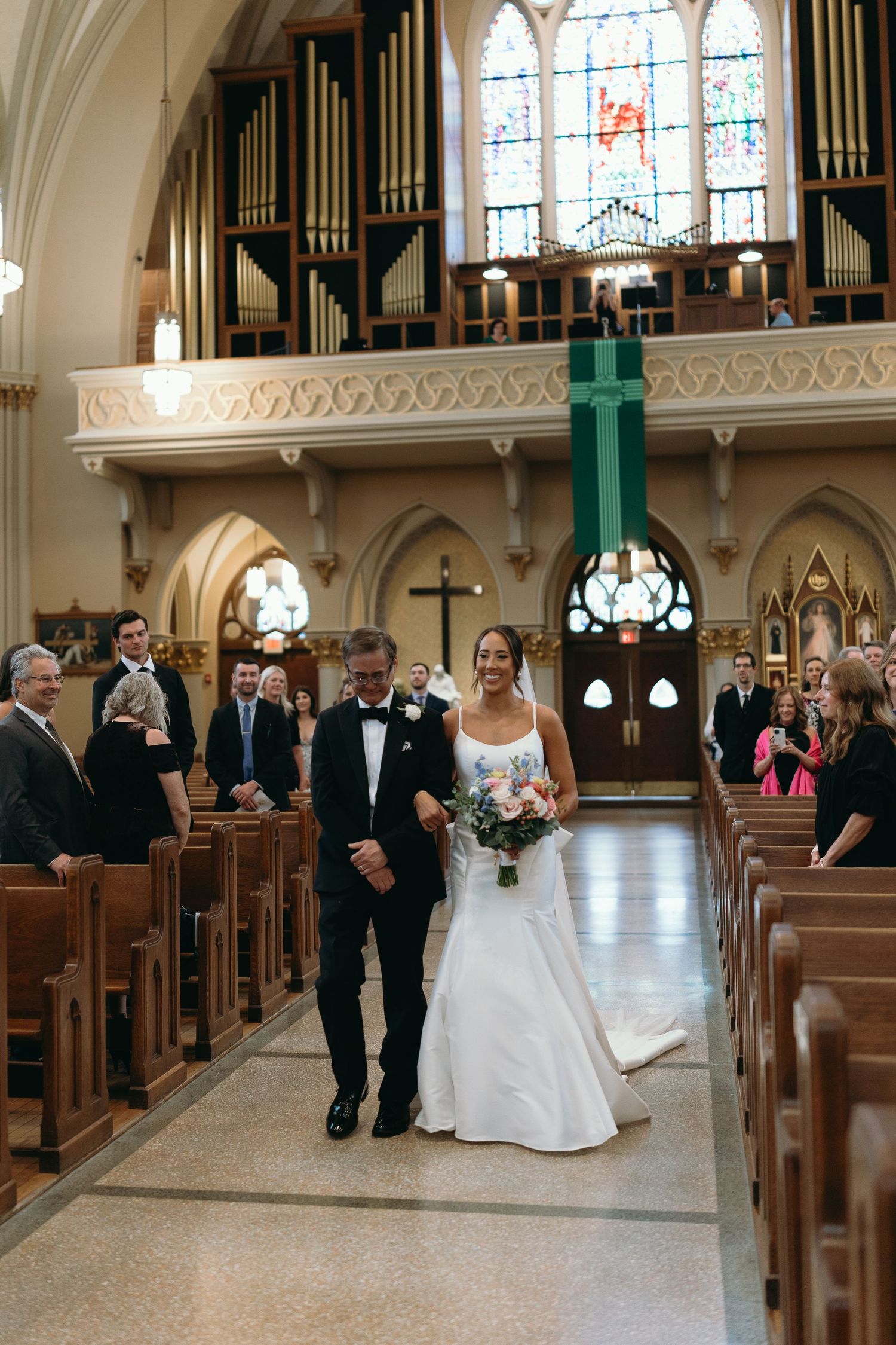 Wedding processional in church aisle with ornate architecture and stained glass windows.