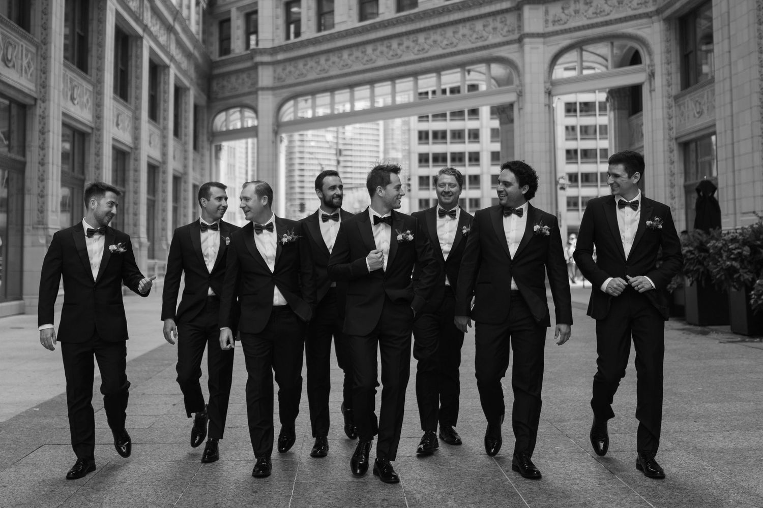 A black and white photograph shows a group of formally dressed men walking in a line down a city street.