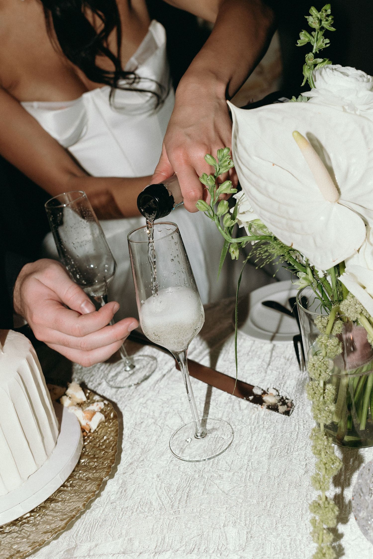 A sequence of hands pouring a creamy white cocktail into elegant champagne glasses on a textured white surface.