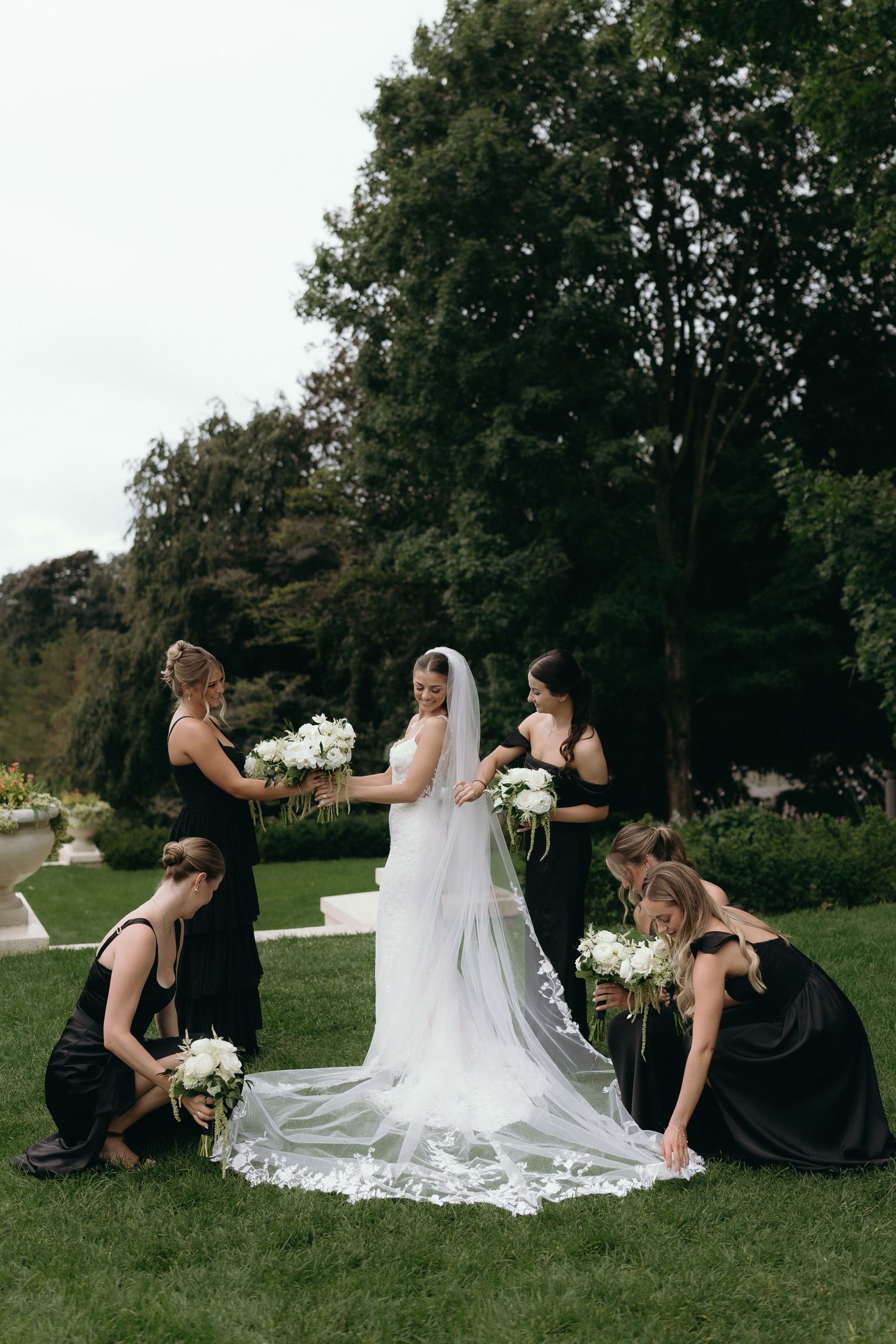 Bride in white gown and veil surrounded by bridesmaids in black dresses arranging her train on green lawn.