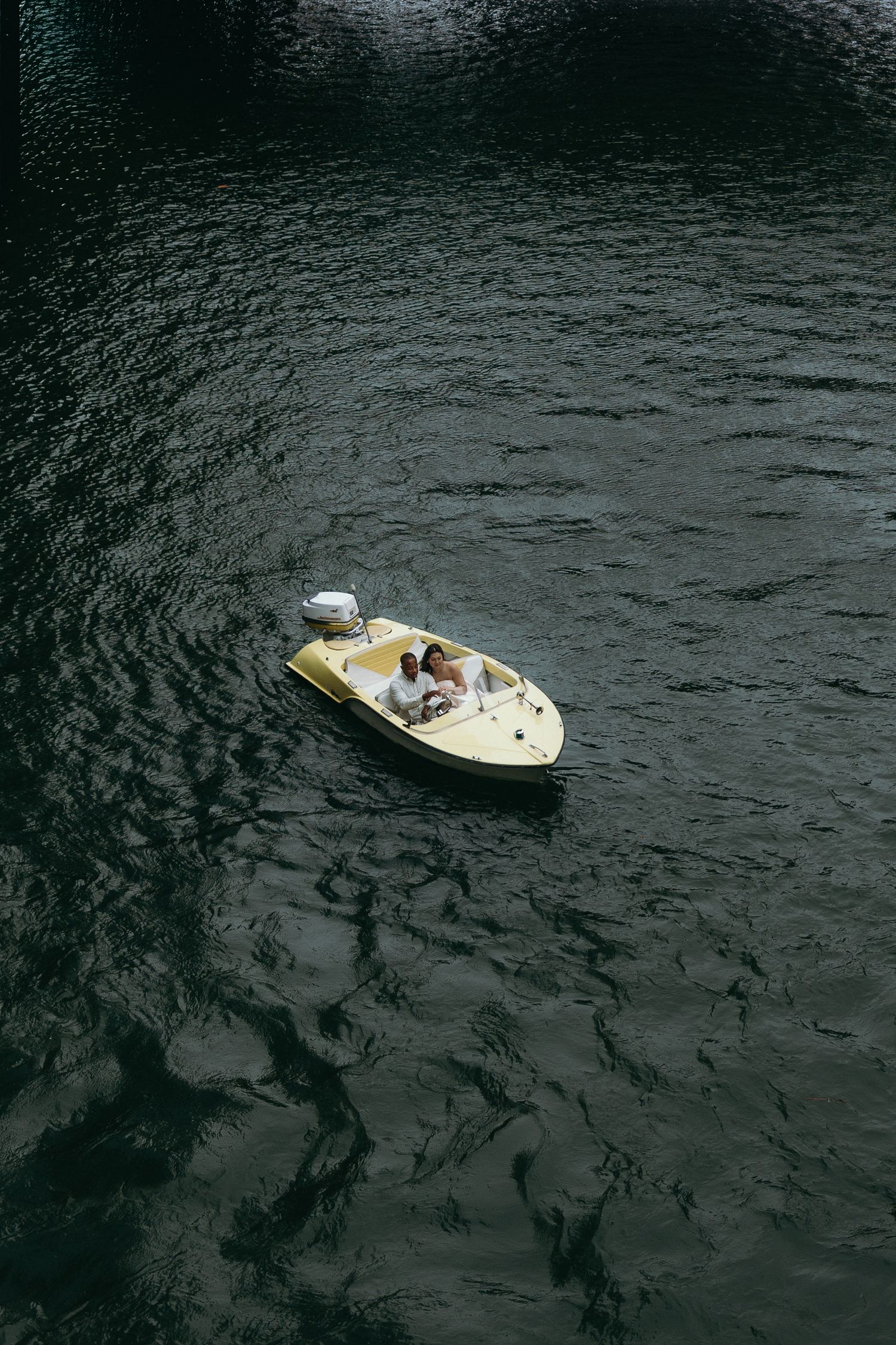 Aerial view of two people relaxing in a small yellow motorboat floating on dark green rippling water.