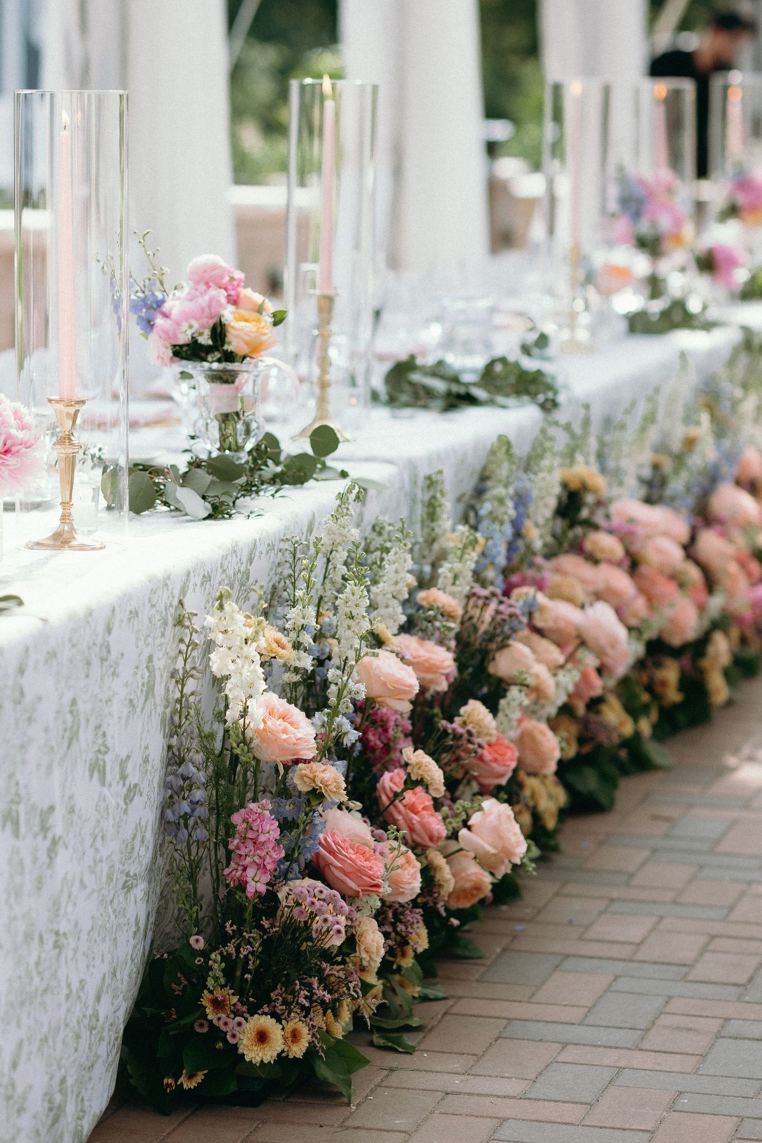 Elegant wedding reception table adorned with lush pink and white floral arrangements, greenery, and tall glass candle holders.