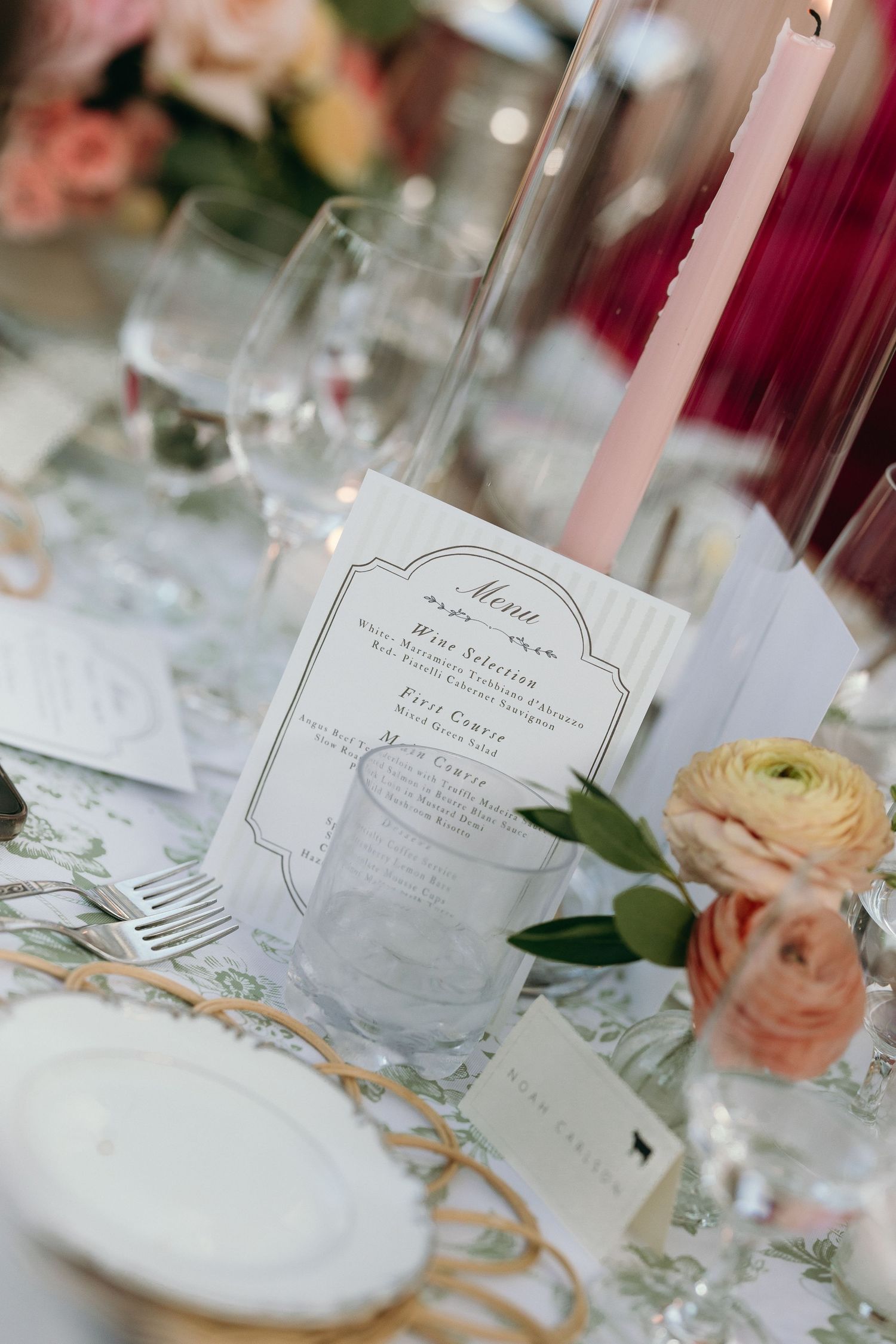 Elegant wedding reception table setting with floral arrangements, pink taper candle, crystal glassware, and a decorative menu card.