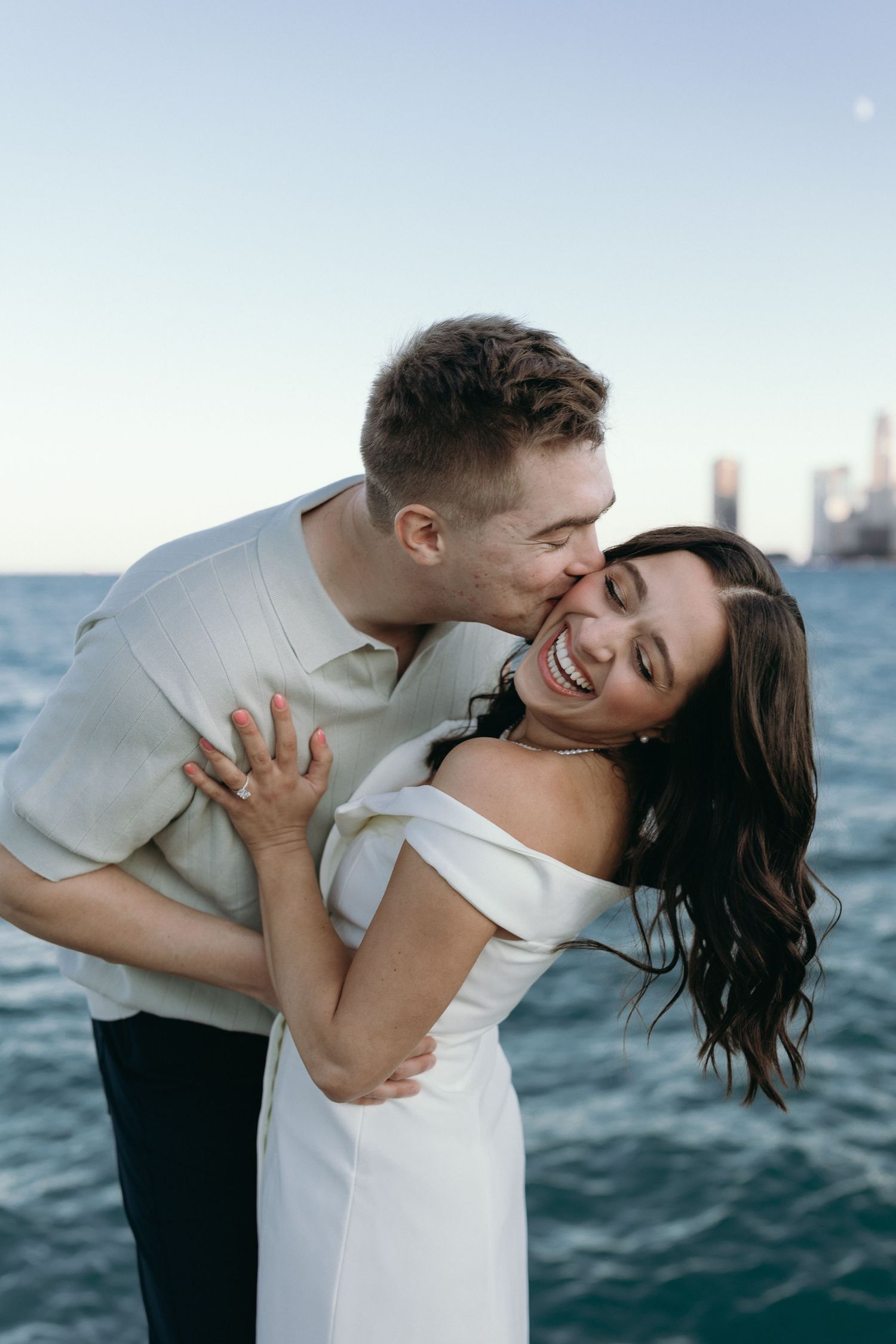 Romantic couple embraces in playful dance poses by the ocean at sunset with dramatic dipping movements.