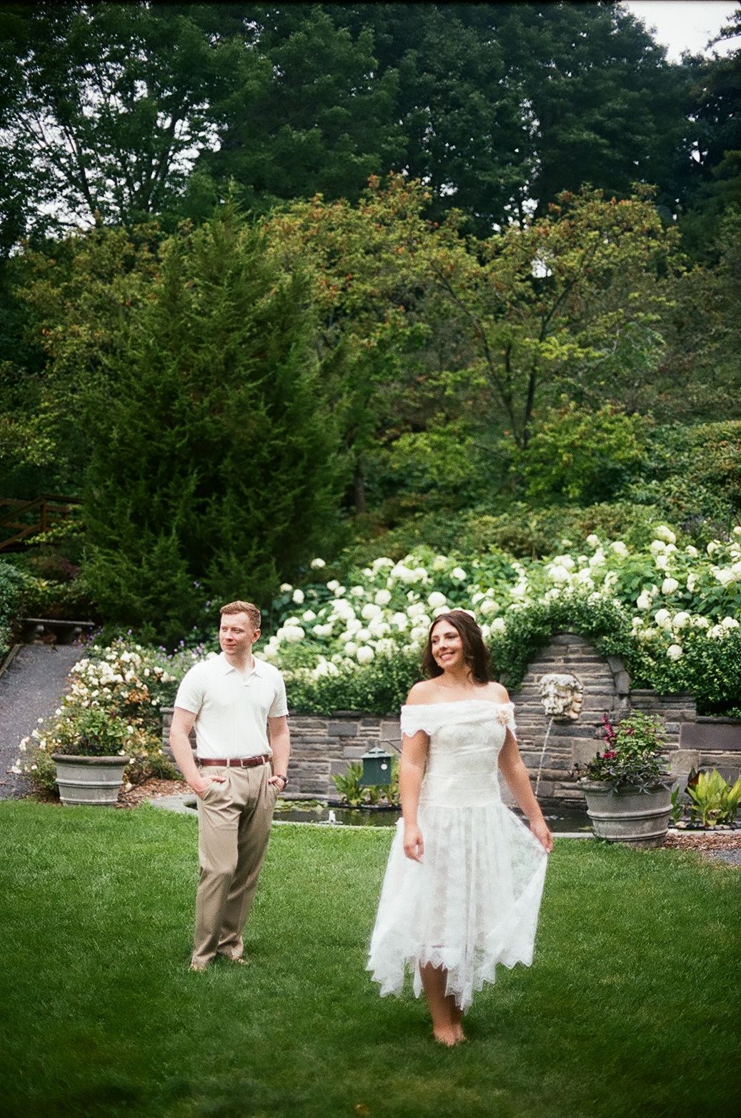 A couple in a garden setting holds up a sheer white dress while walking on grass near flowering bushes and stone retaining walls.