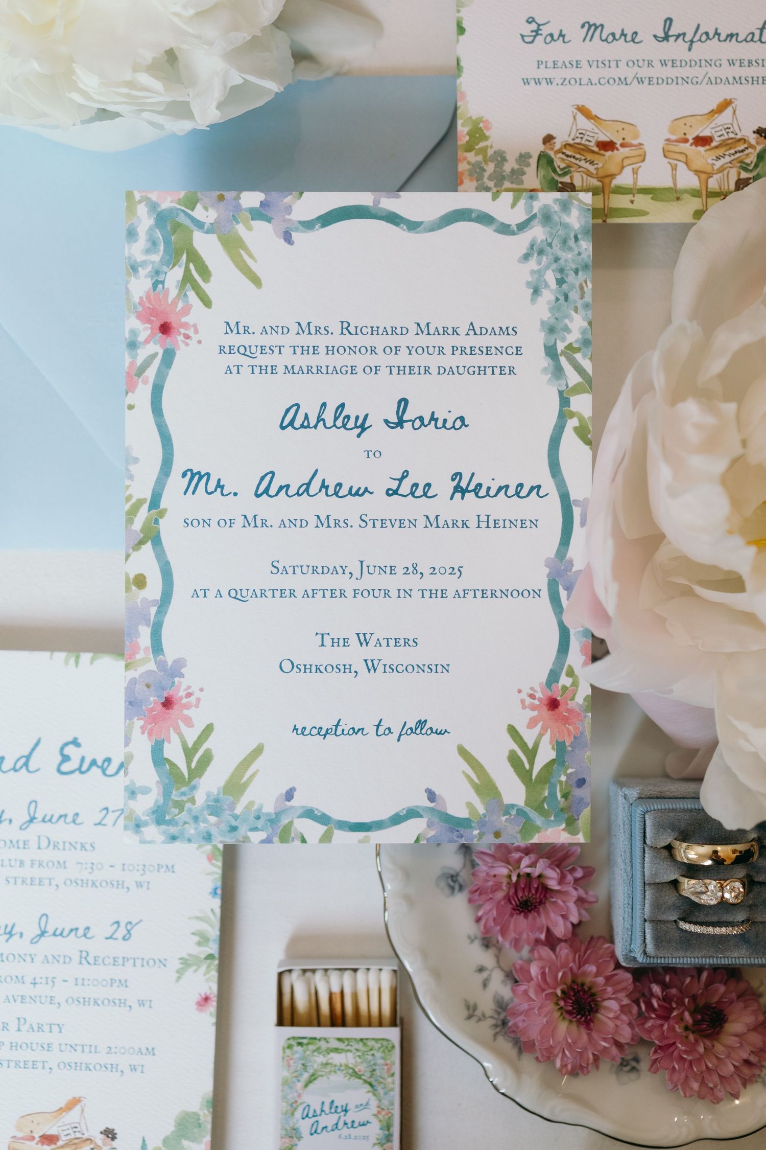 Wedding invitation with floral border design in blue and pink watercolors alongside rings and dried flowers.