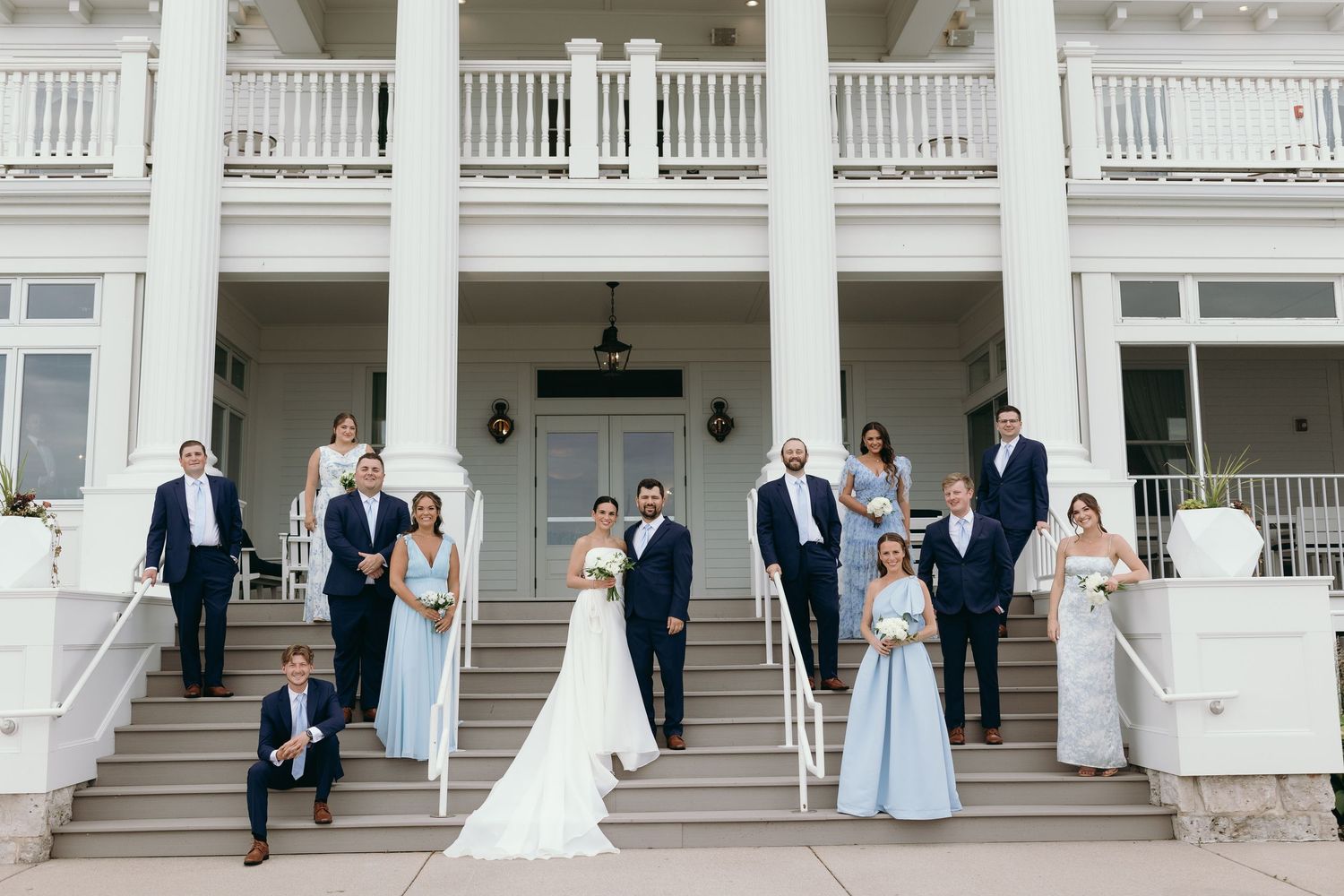 Wedding party in navy and light blue attire poses elegantly on grand white mansion steps during formal celebration.