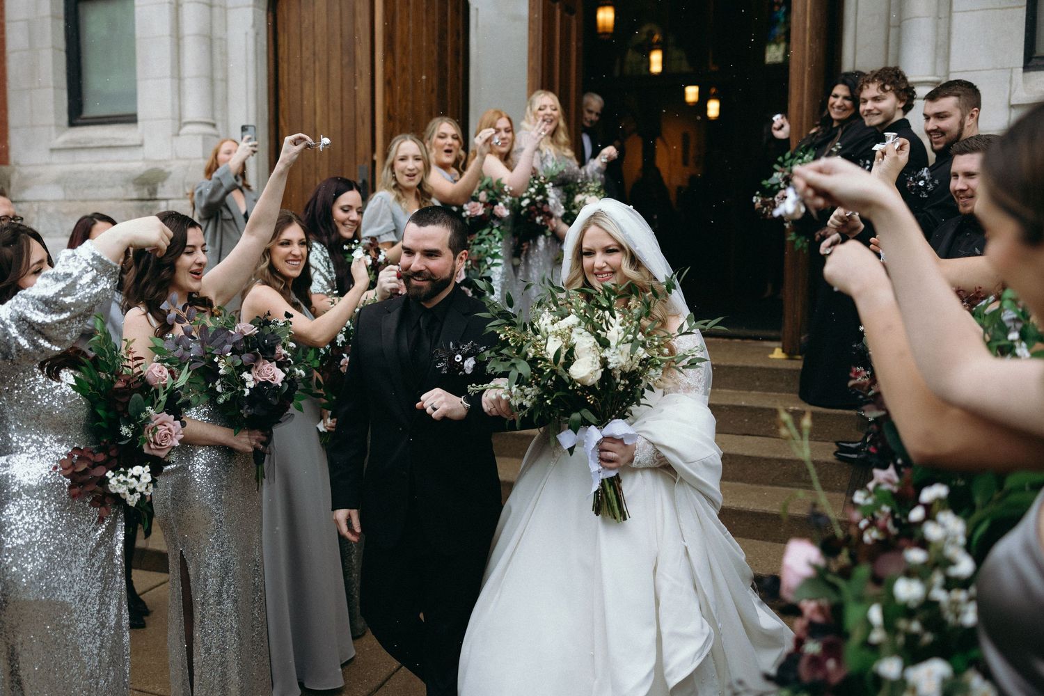 Joyful wedding party celebrates with bouquets on church steps after ceremony.